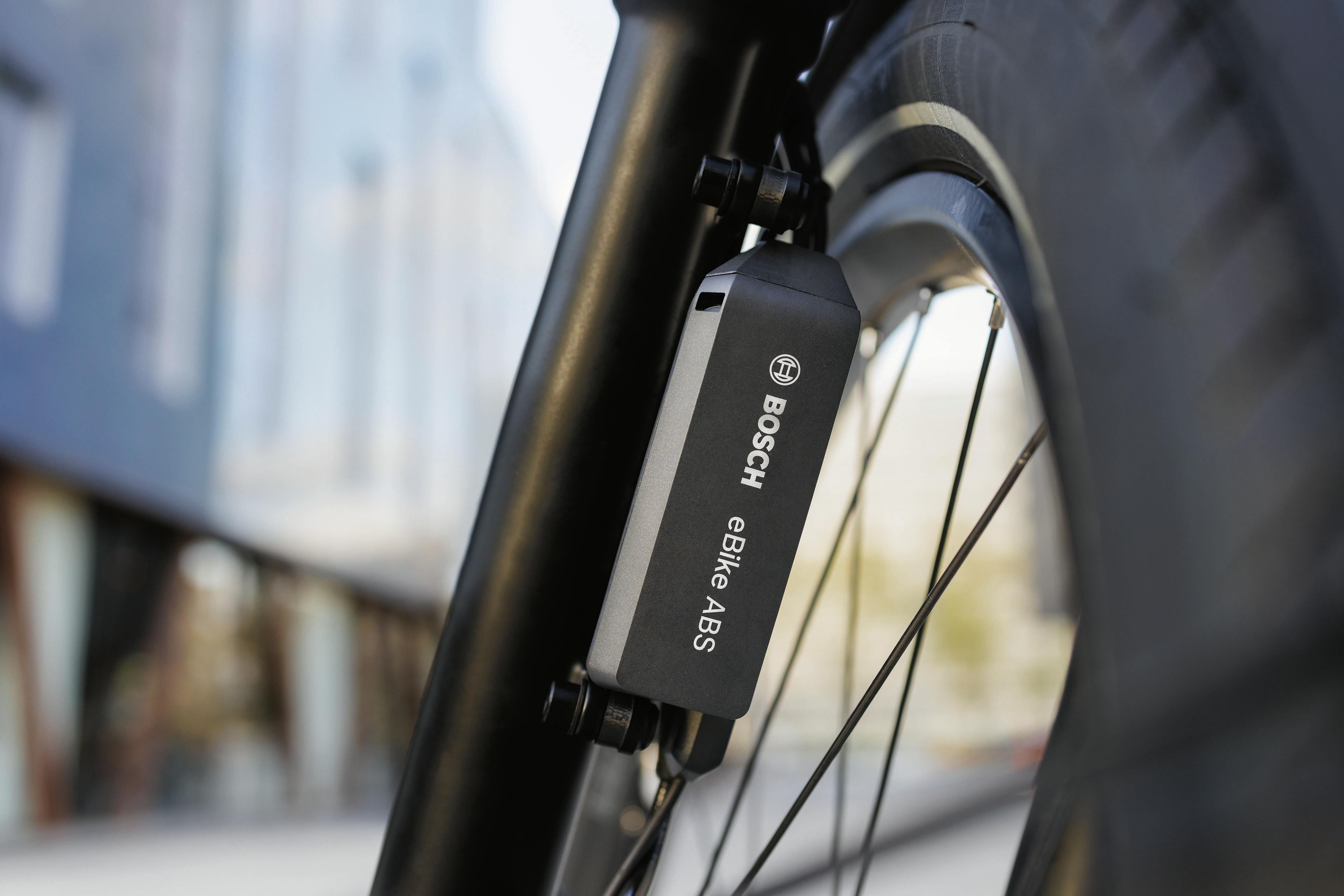 Nahaufnahme eines Bosch eBike-ABS-Sensors, der an der Vorderradgabel eines Fahrrads montiert ist. Im Hintergrund sind moderne Gebäude zu sehen.