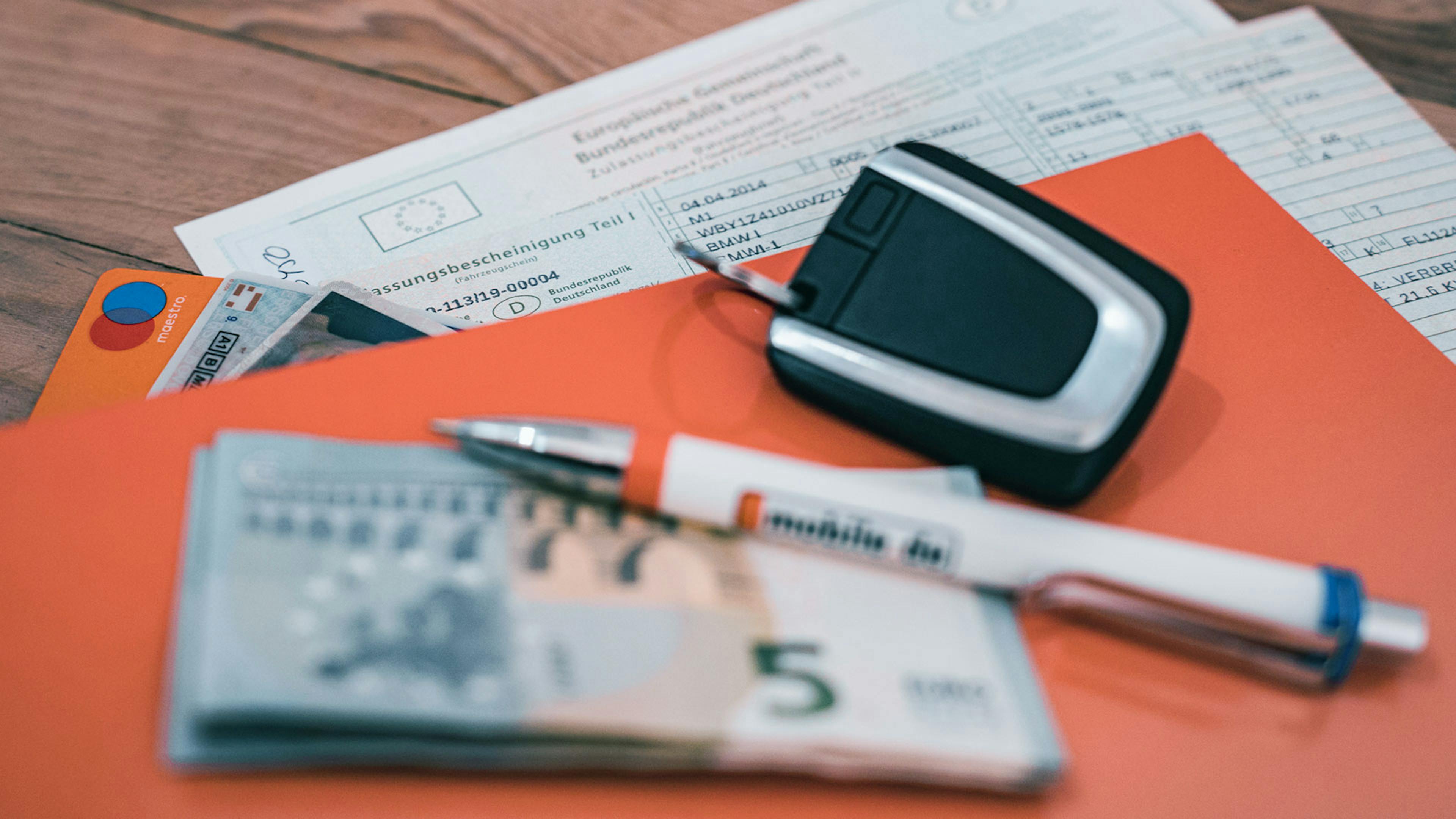 Eine orangefarbene Infomappe mit einem Fahrzeugbrief und weiteren Unterlagen liegt auf einem Holztisch, darauf ein Autoschlüssel, ein Kugelschreiber und diverse Geldscheine.