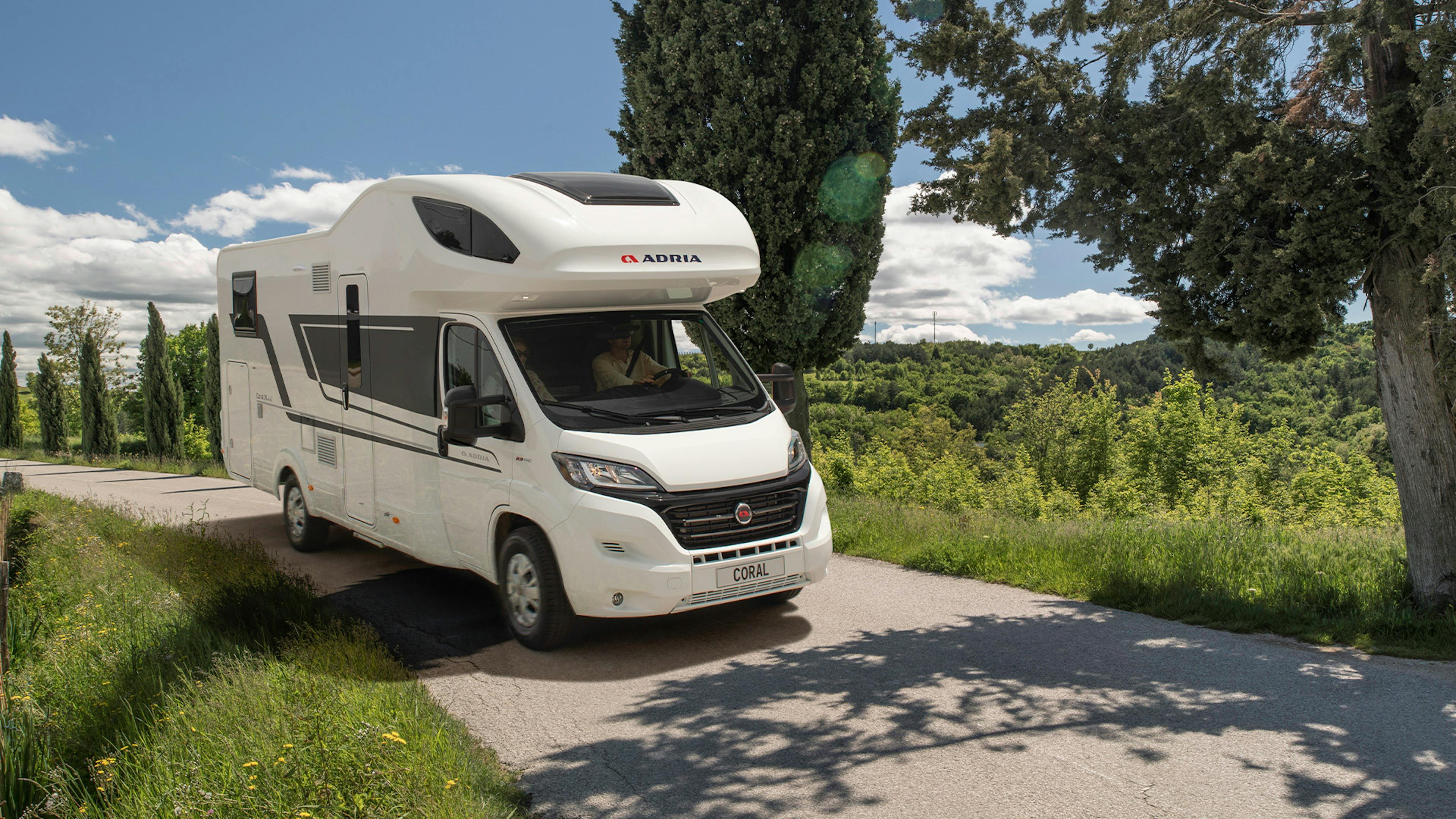 Ein Coral XL Wohnmobil von Adria Mobile fährt eine Straße entlang durch eine mediterrane Landschaft.