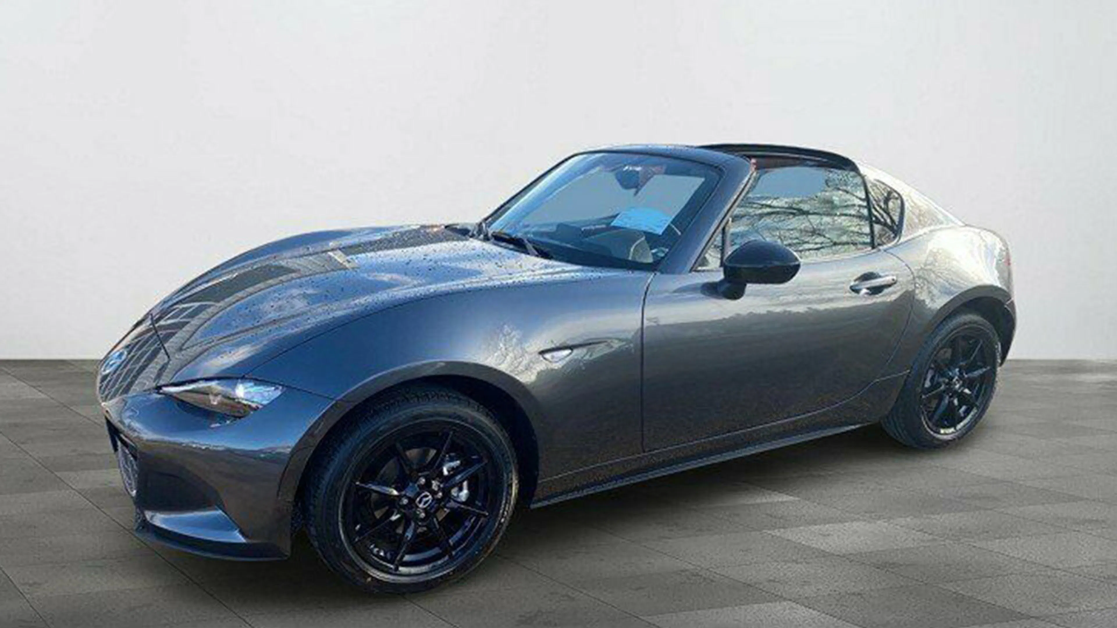 Grauer Sportwagen mit Hardtop, schwarzer Felgen und aerodynamischem Design, seitlich fotografiert auf grauem Boden vor einer weißen Wand.