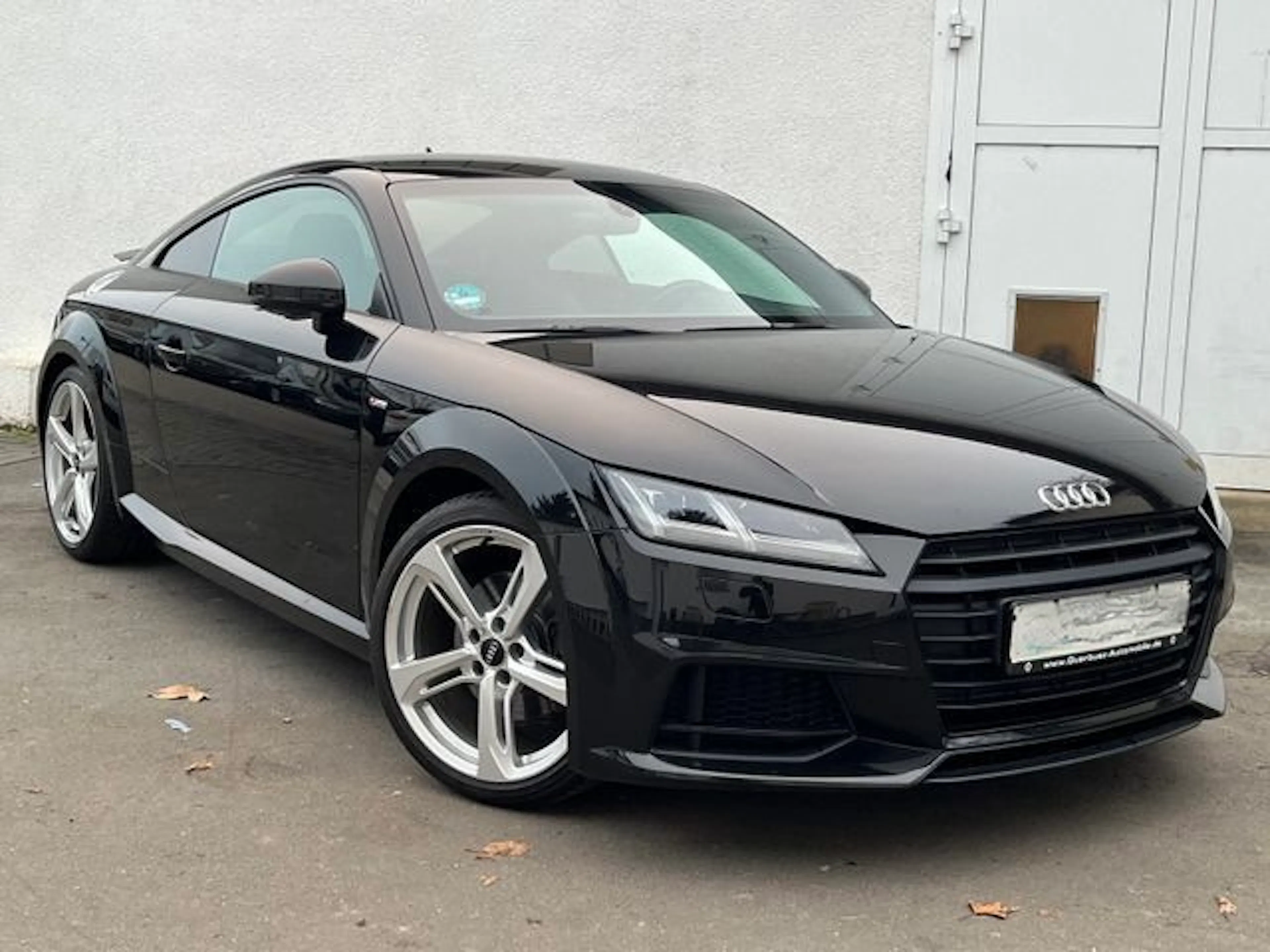 Schwarzer Audi TT Coupé steht auf einem betonierten Platz vor einer weißen Wand, schräg von vorn fotografiert. Moderne LED-Scheinwerfer und Alufelgen.