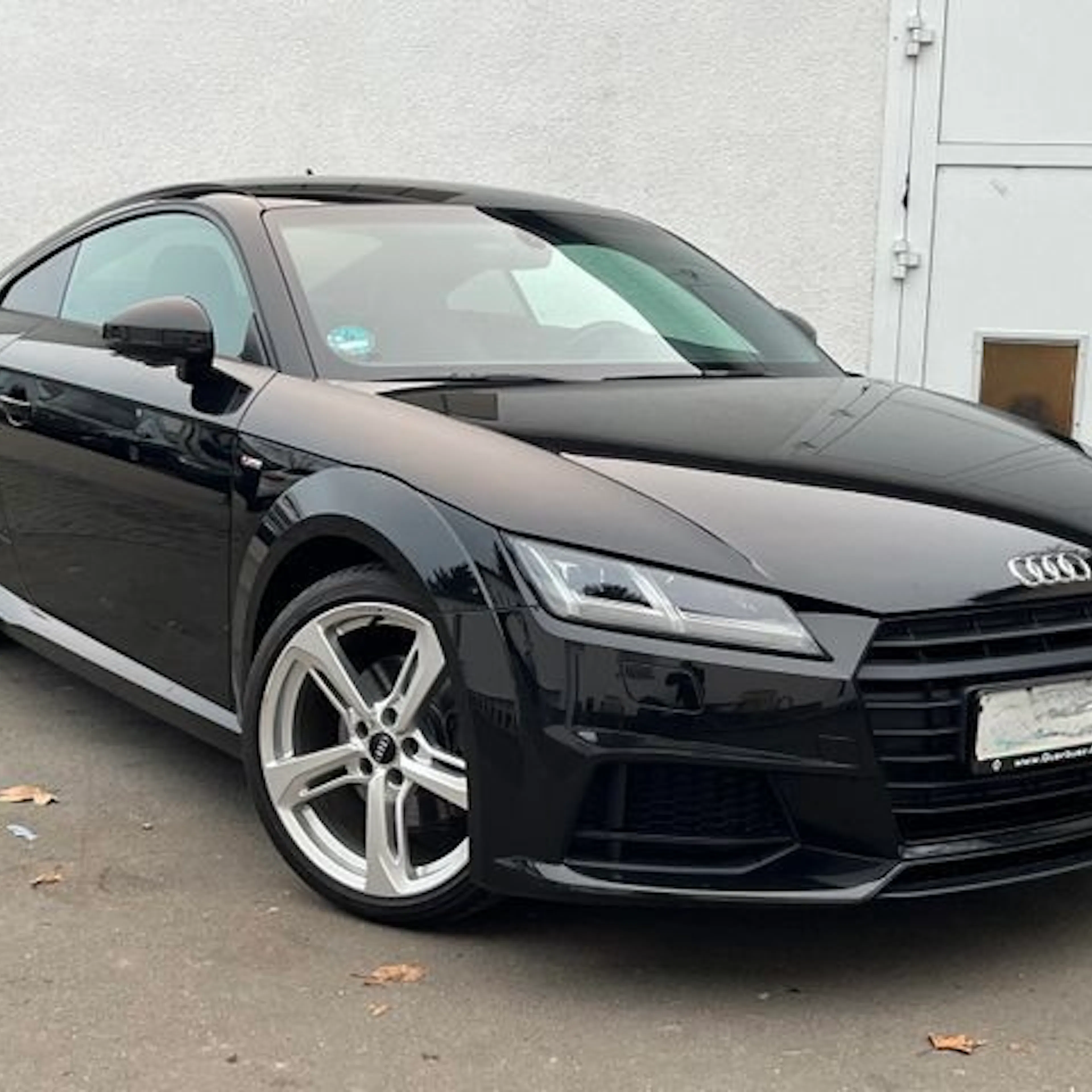 Schwarzer Audi TT Coupé steht auf einem betonierten Platz vor einer weißen Wand, schräg von vorn fotografiert. Moderne LED-Scheinwerfer und Alufelgen.