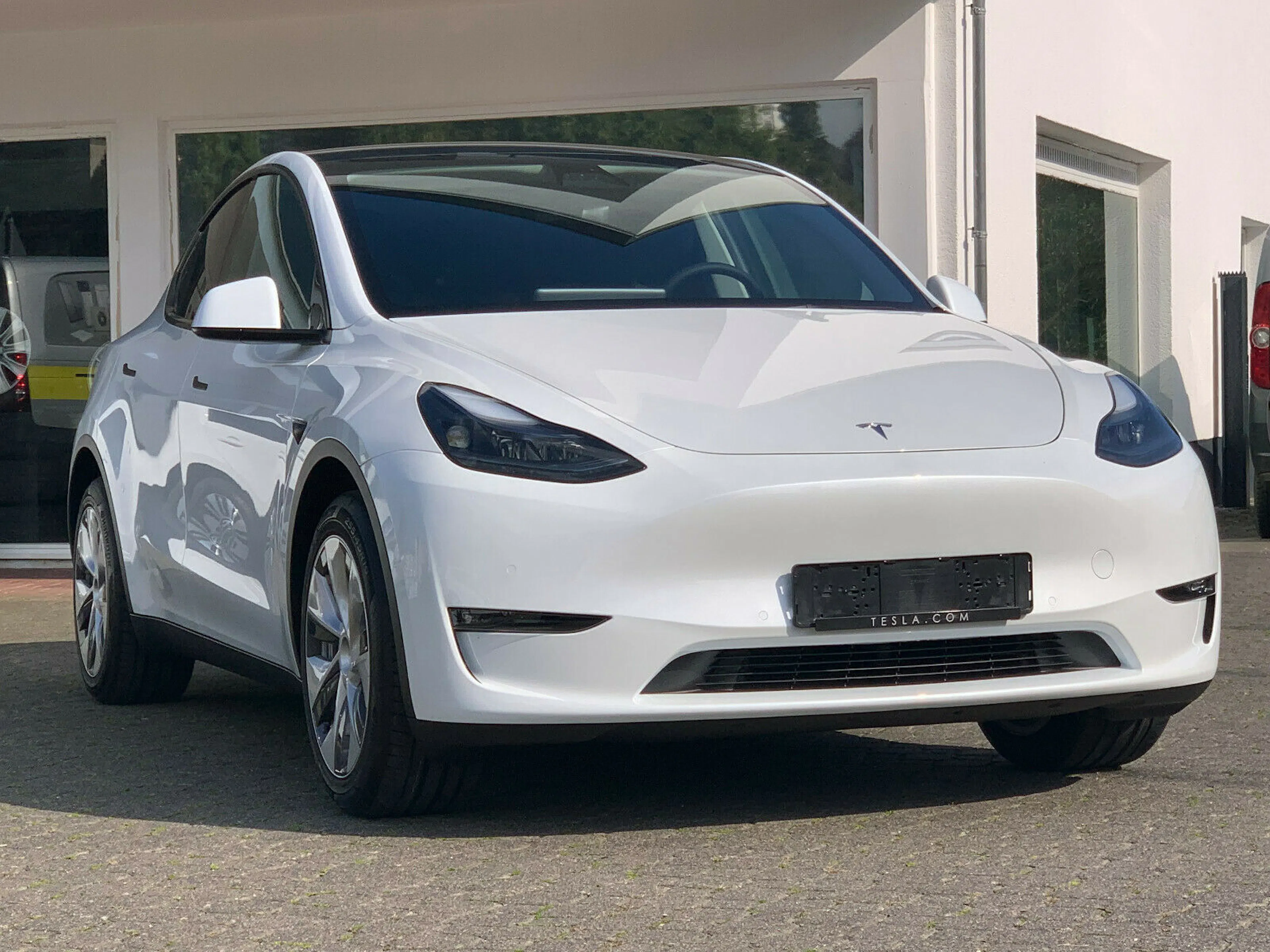 Weißes Elektroauto, Tesla Model Y, steht auf einem Parkplatz vor einem Gebäude mit reflektierenden Fenstern.