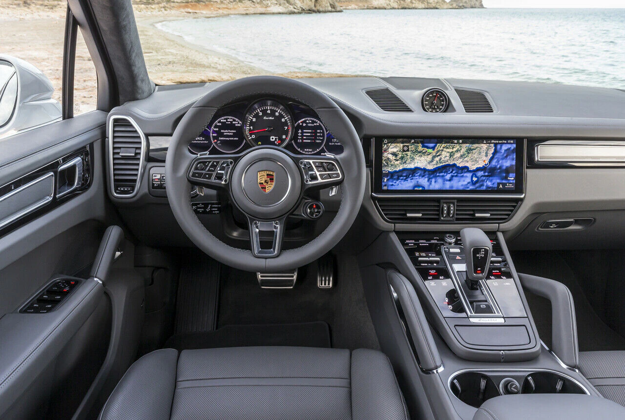 Innenraum eines modernen Autos mit Lederlenkrad, digitalen Armaturen, großem Touchscreen und Getriebe; Strand im Hintergrund sichtbar.