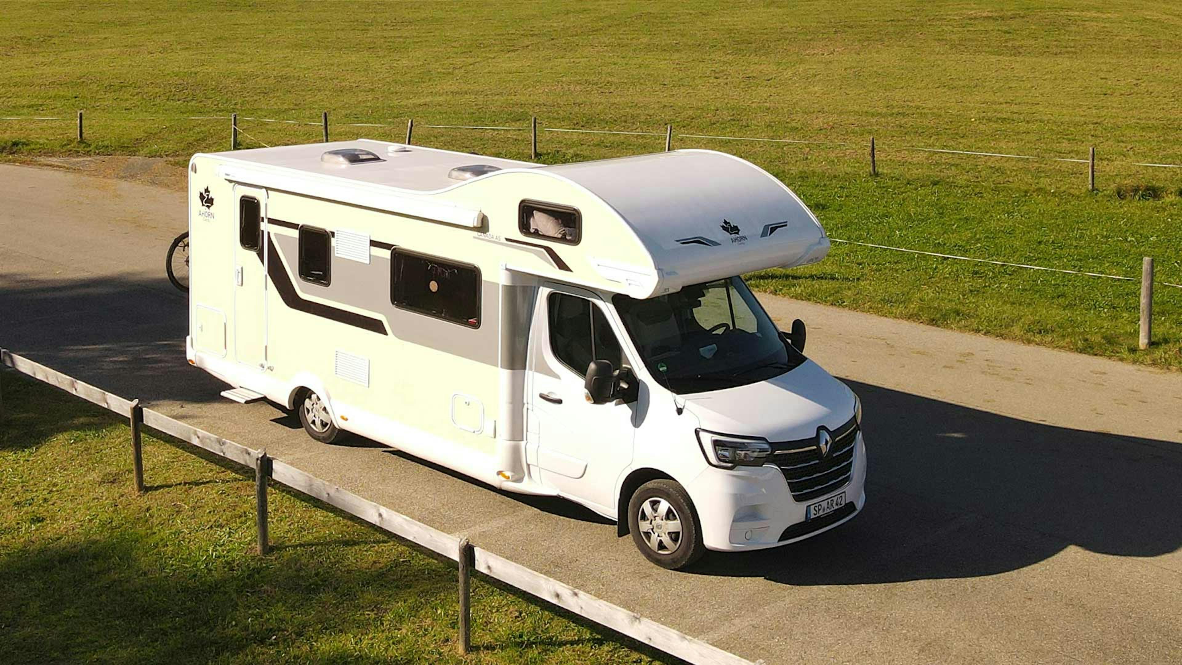 Ein weißes Ahorn Camp Canada AS Wohnmobil mit Alkoven fährt eine Landstraße entlang.