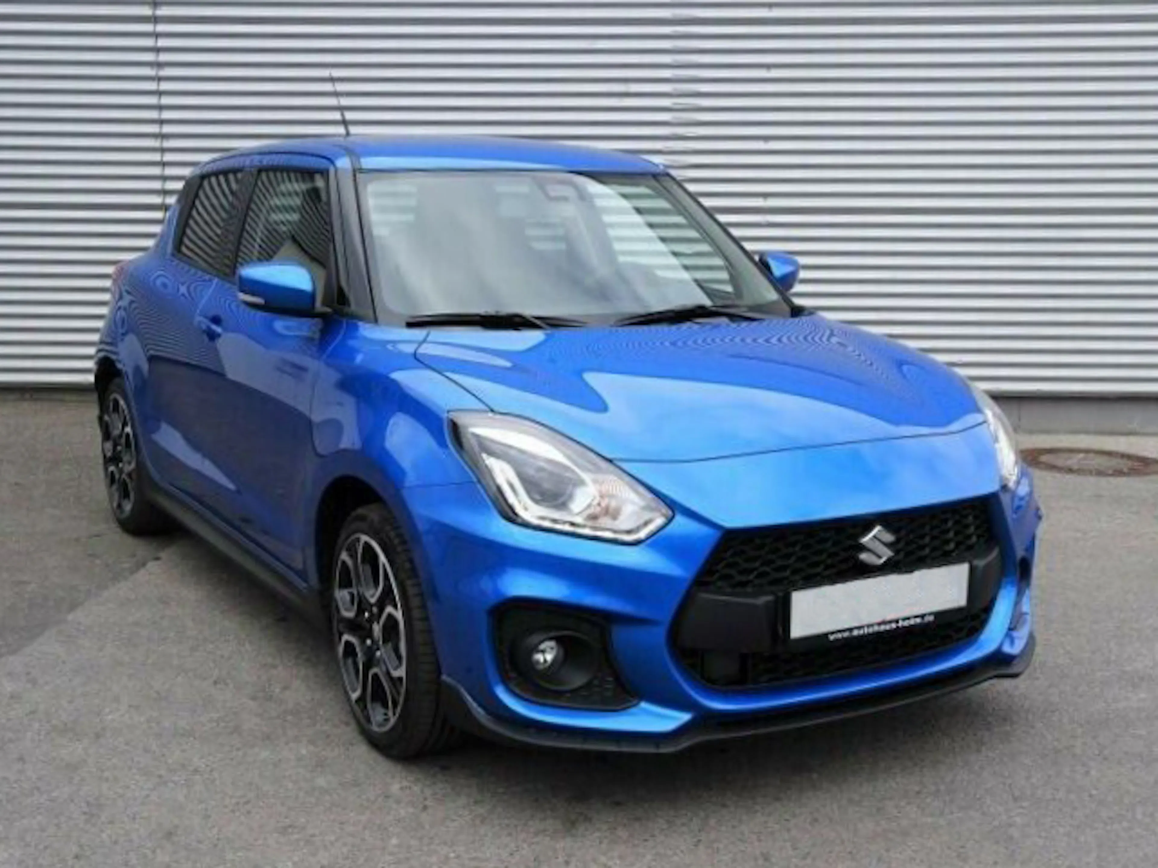 Blauer Suzuki Swift steht vor einer silbernen Wellblechwand, leicht von rechts vorn gesehen, auf grauem Asphalt.