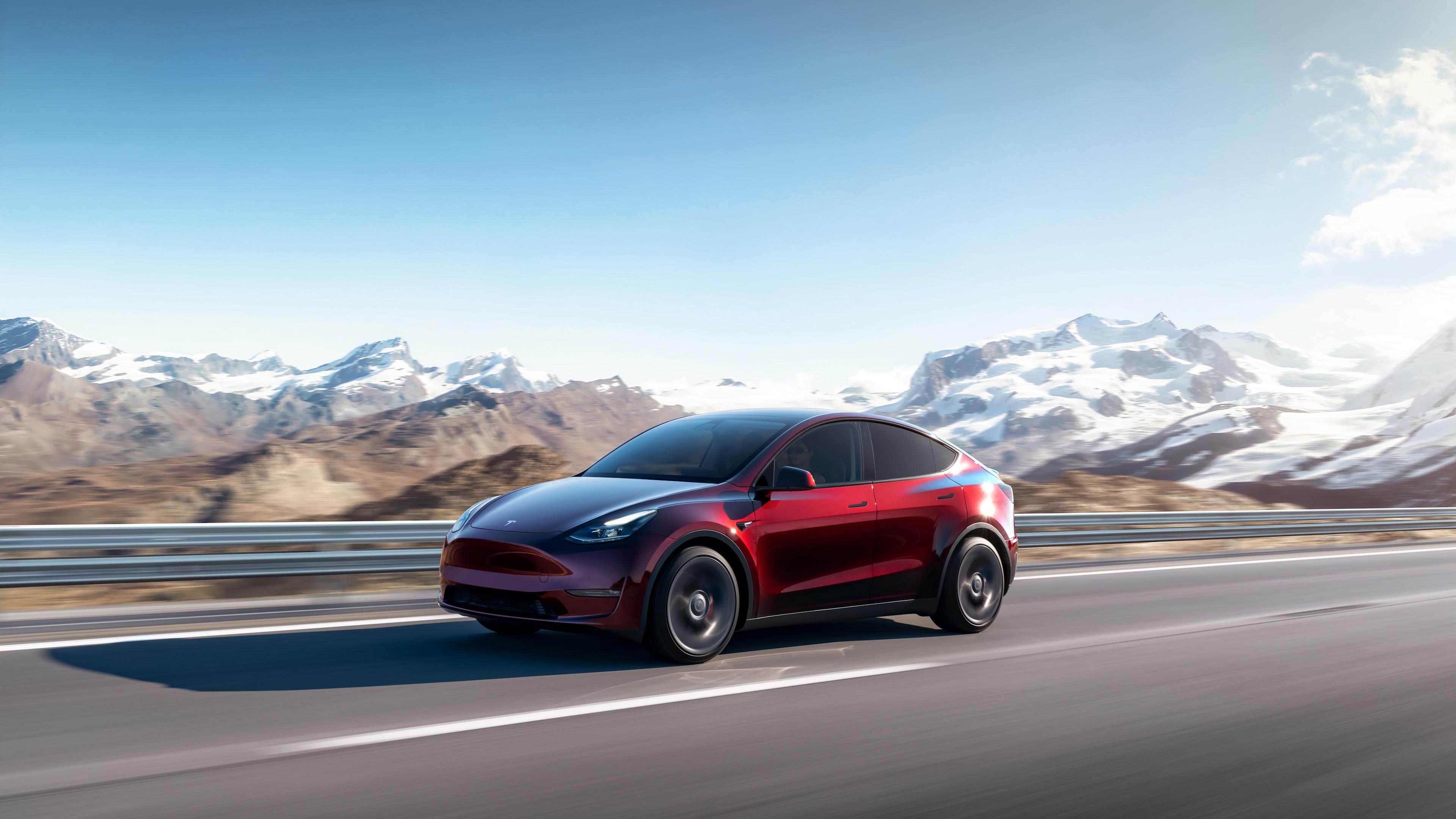 A red Tesla Model Y is driving on a country road with a mountain range in the background.