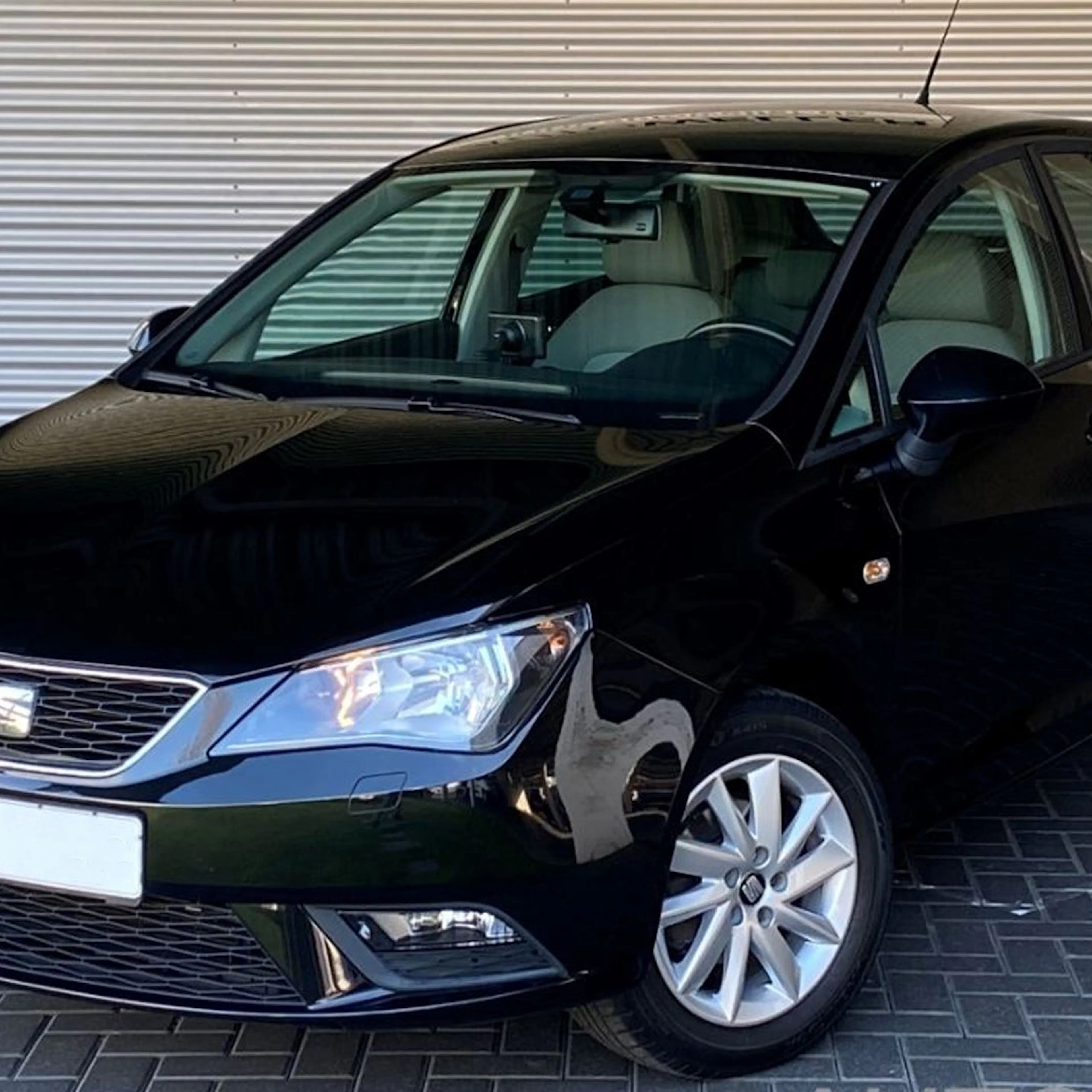 Schwarzer Seat-Ibiza-Kleinwagen, schräg von vorn fotografiert, vor einer Metallwand auf gepflastertem Untergrund geparkt.