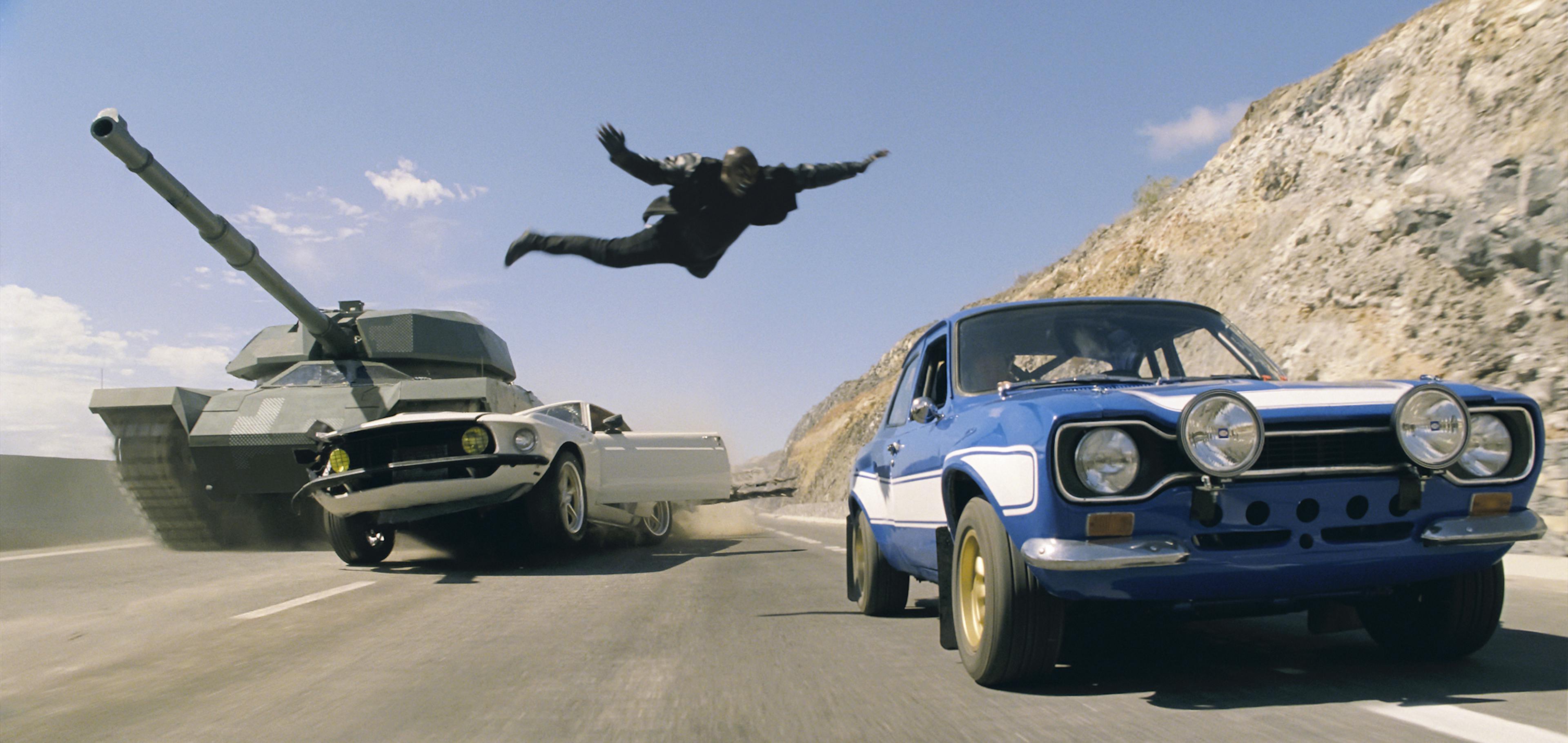 Ein Ford Escort RS 2000 fährt auf einer Schnellstraße, während im Hintergrund ein Ford Mustang von einem Panzer zermalmt wird.