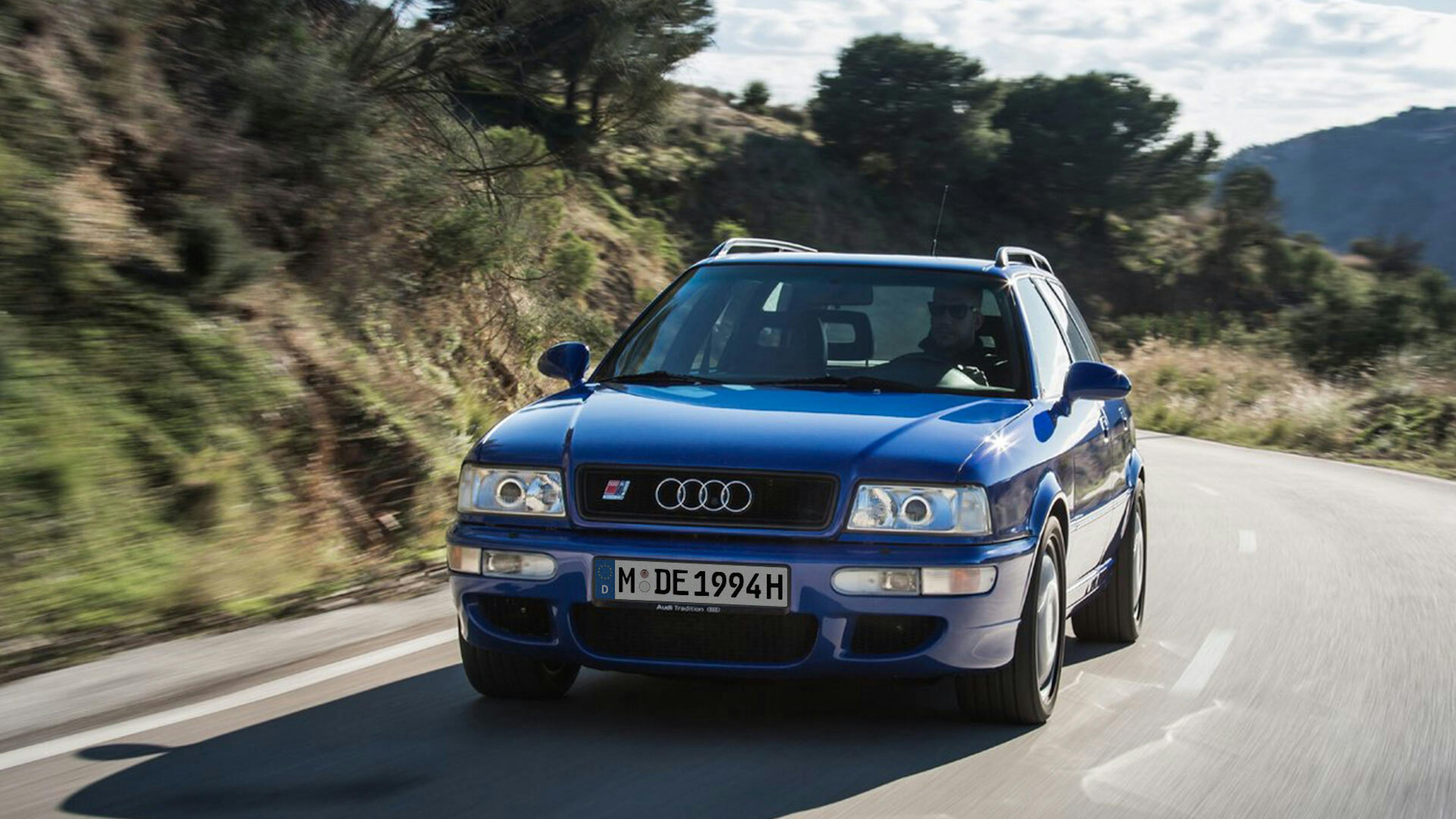 Blauer Audi SUV Oldtimer fährt auf kurviger Landstraße bei sonnigem Wetter, umgeben von Hügeln und Bäumen.