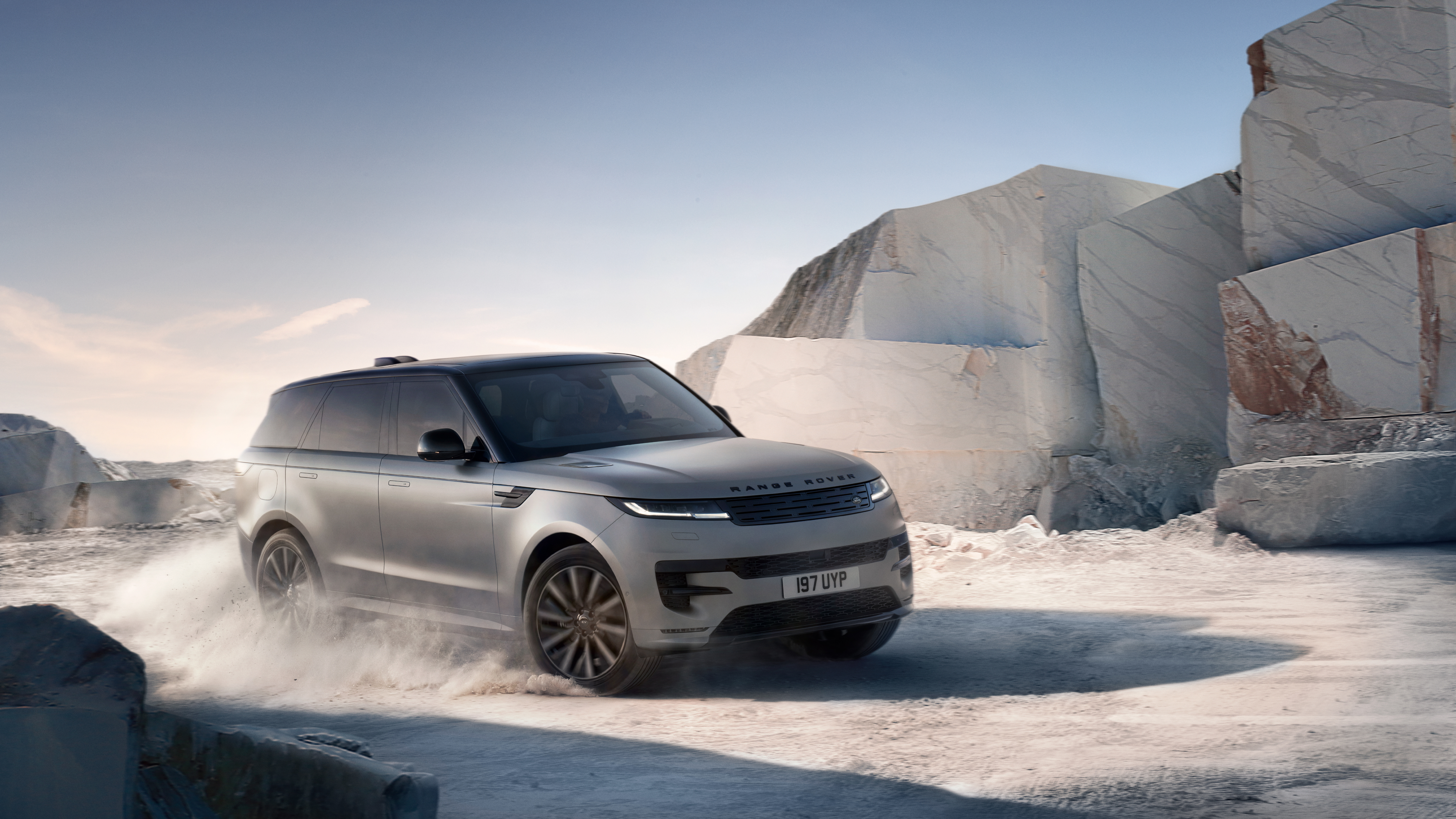 Grauer Range Rover Sport Electric wirbelt Staub auf einem Schotterweg vor hellen Marmorblöcken auf.