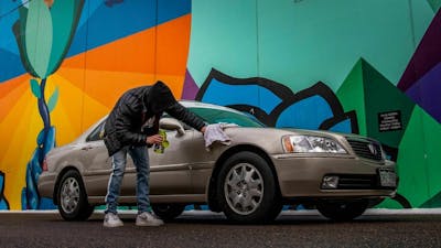 Ein junger Mann poliert einen grauen Pkw, der vor einer Wand mit Graffiti steht.