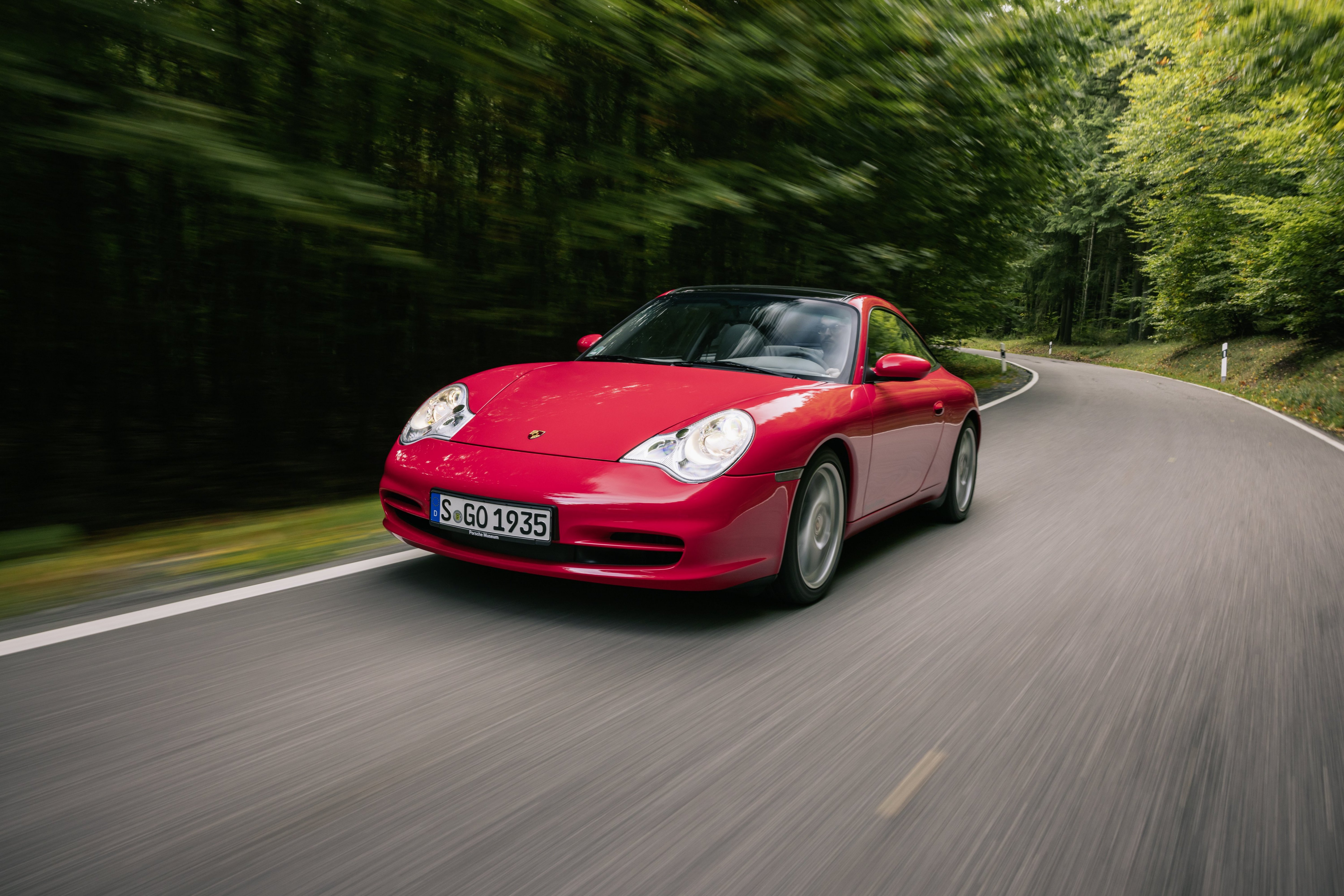 Roter Porsche 911 der Baureihe 996.2 fährt dynamisch auf einer Landstraße, Frontansicht mit Bewegungsunschärfe im Hintergrund.