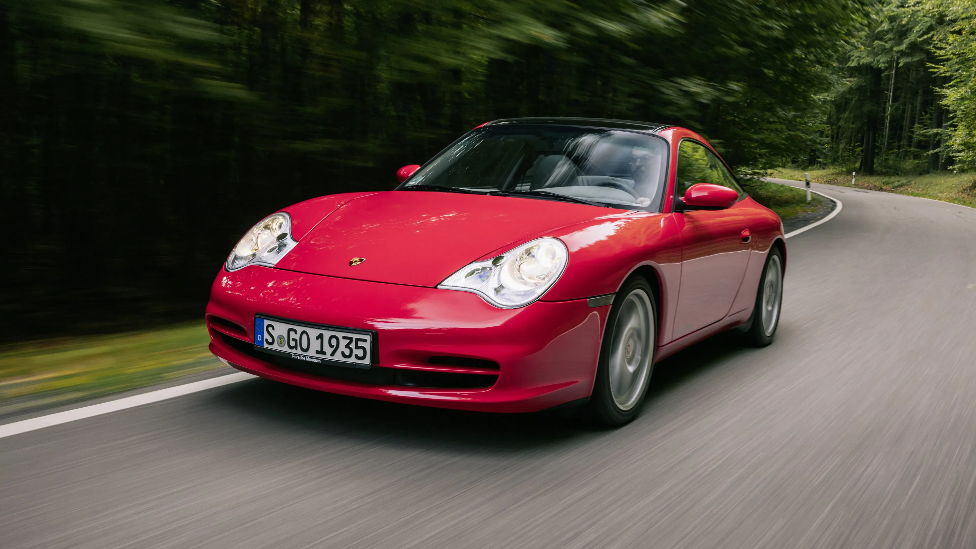 Roter Porsche 911 der Baureihe 996.2 fährt dynamisch auf einer Landstraße, Frontansicht mit Bewegungsunschärfe im Hintergrund.