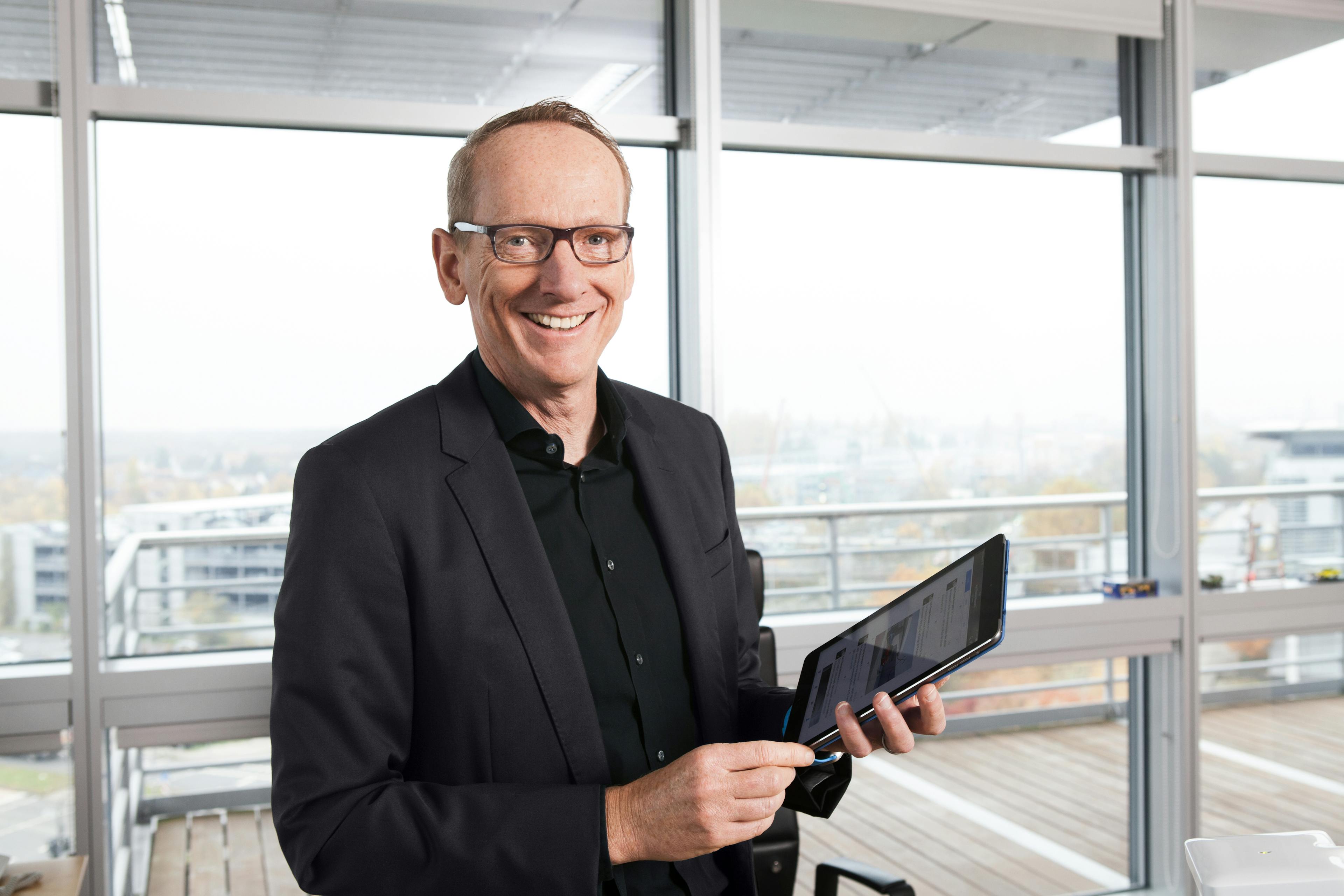 Ein lächelnder Mann im Anzug hält ein Tablet in einem modernen Büro mit großen Fenstern und Blick auf eine Stadtlandschaft.