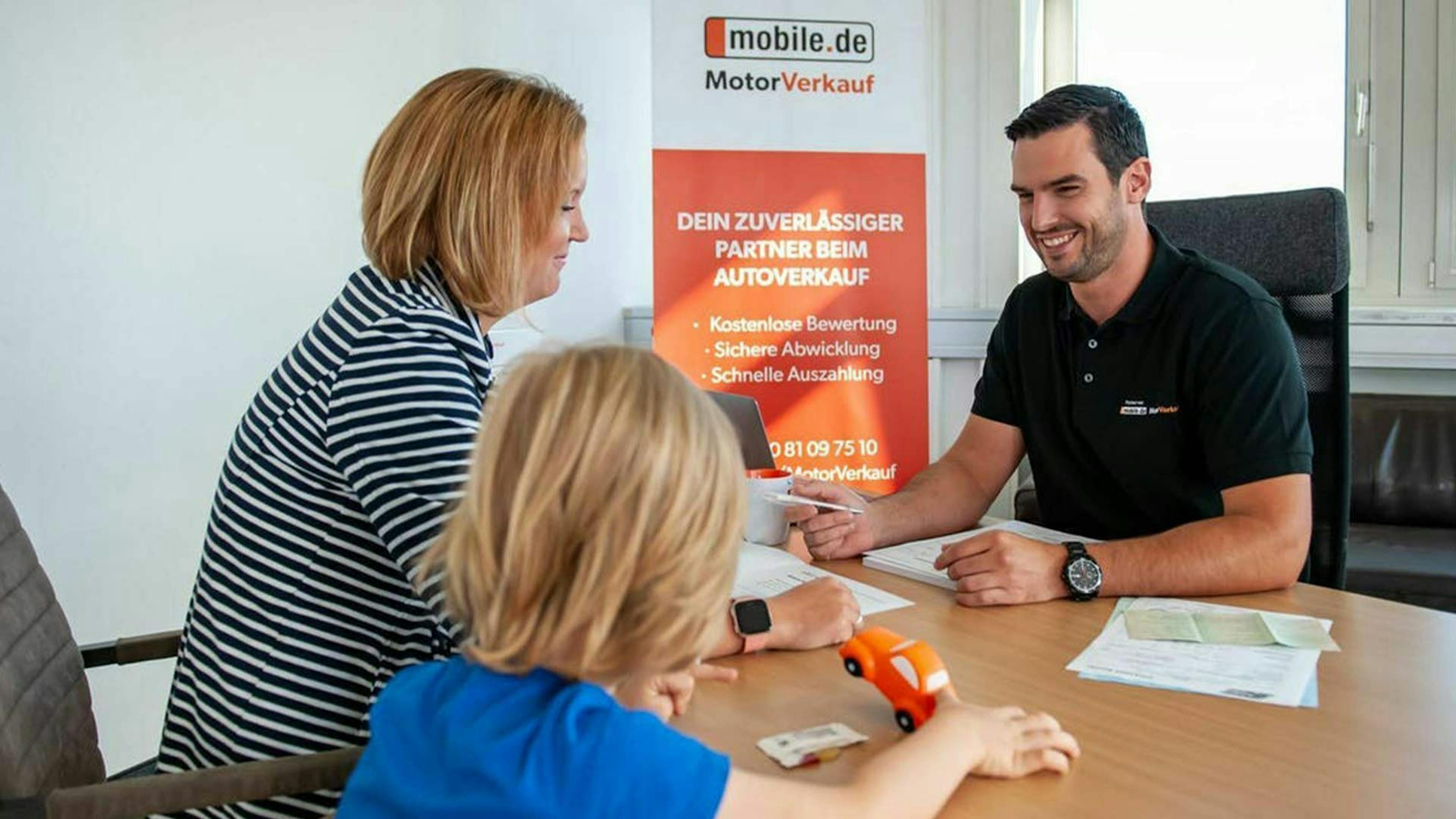 Eine Frau und ein Kind sitzen an einem Tisch und lassen sich zum Thema Autokauf-Preisverhandlung von einem mobile.de Mitarbeiter beraten.