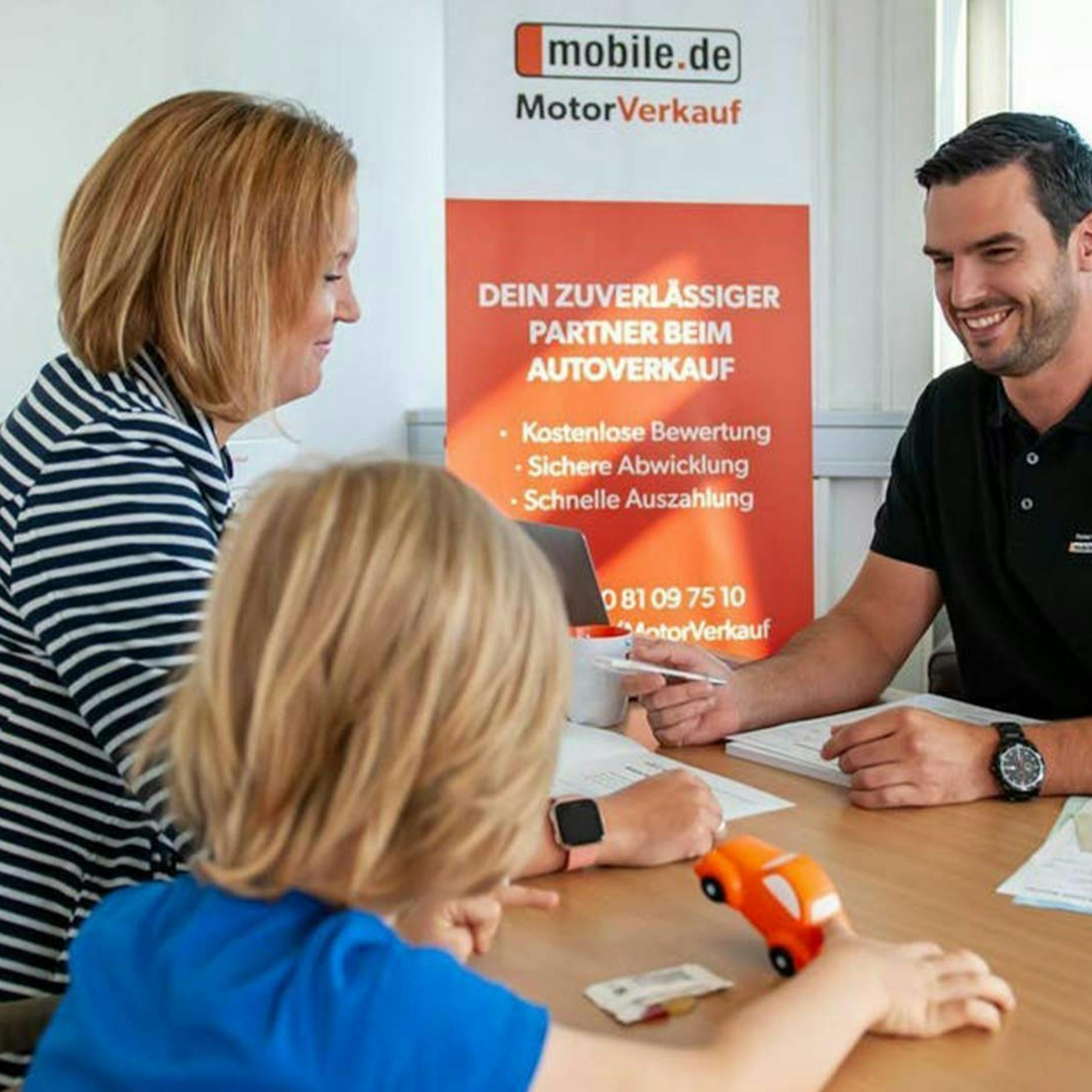 Eine Frau und ein Kind sitzen an einem Tisch und lassen sich zum Thema Autokauf-Preisverhandlung von einem mobile.de Mitarbeiter beraten.