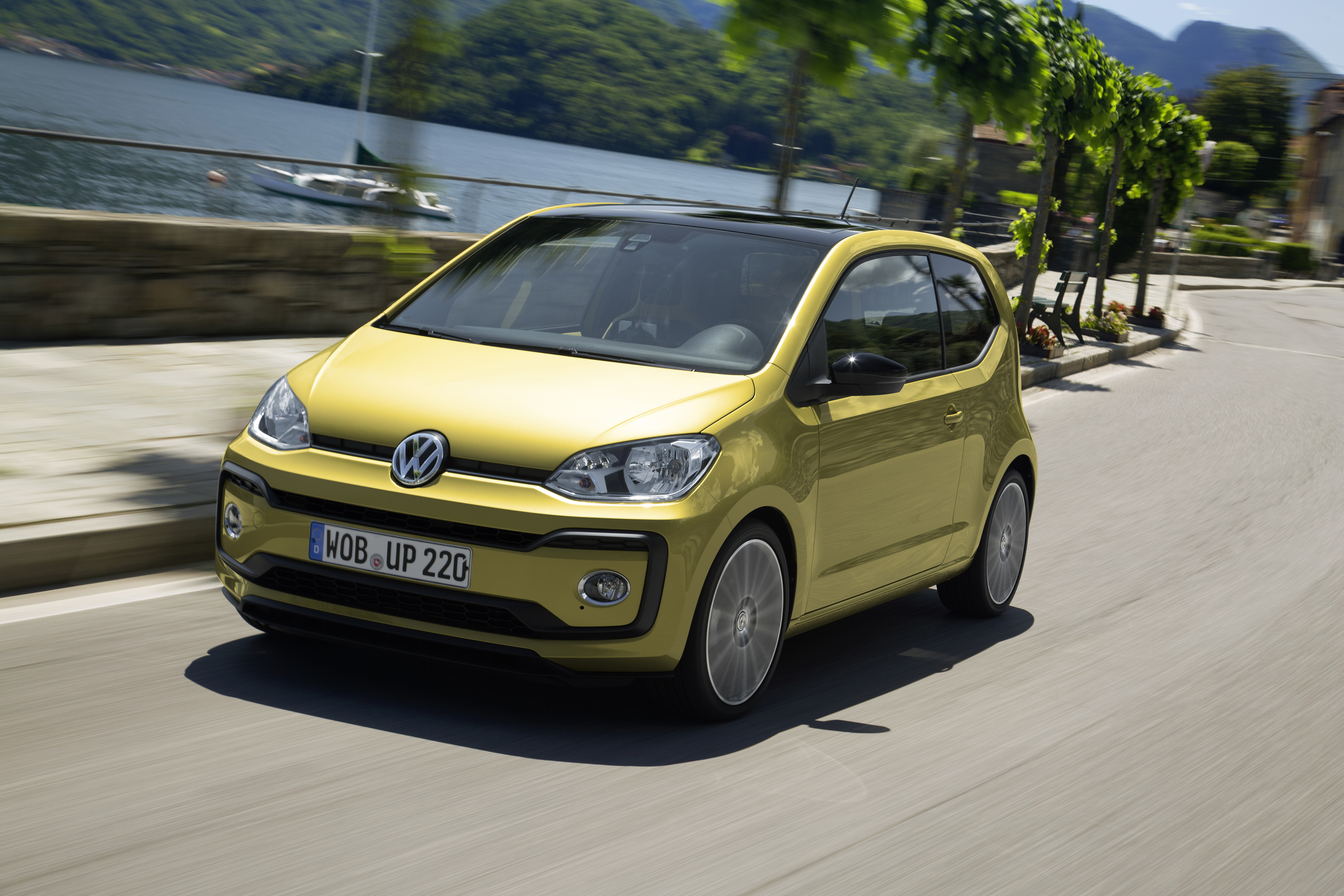 Goldgelber VW up! als Dreitürer fährt auf einer Uferstraße, im Hintergrund sind ein See und bewaldete Berge zu sehen.
