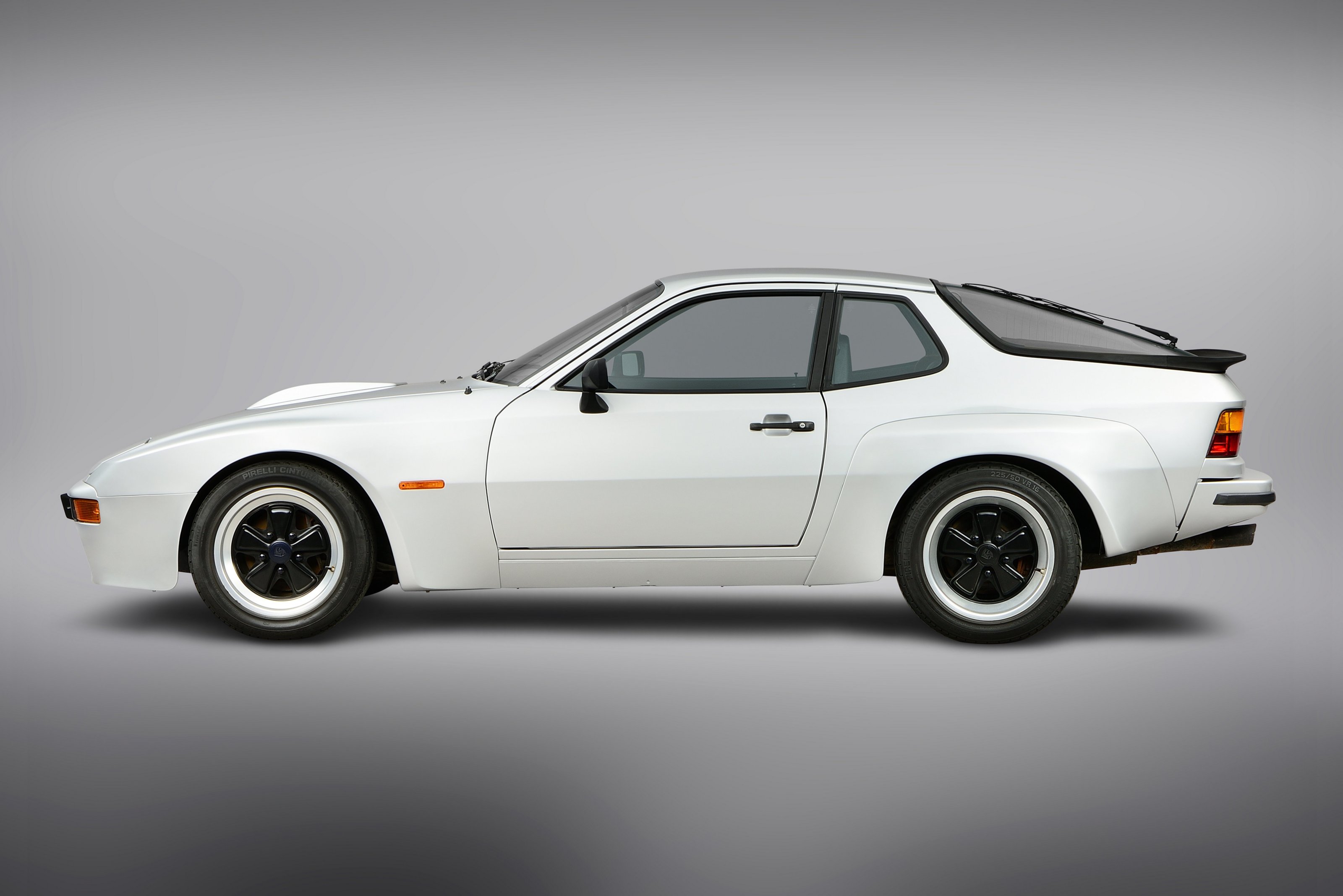 Weißer Porsche 924 steht seitlich vor neutralem, grauem Hintergrund, Klappscheinwerfer eingeklappt.