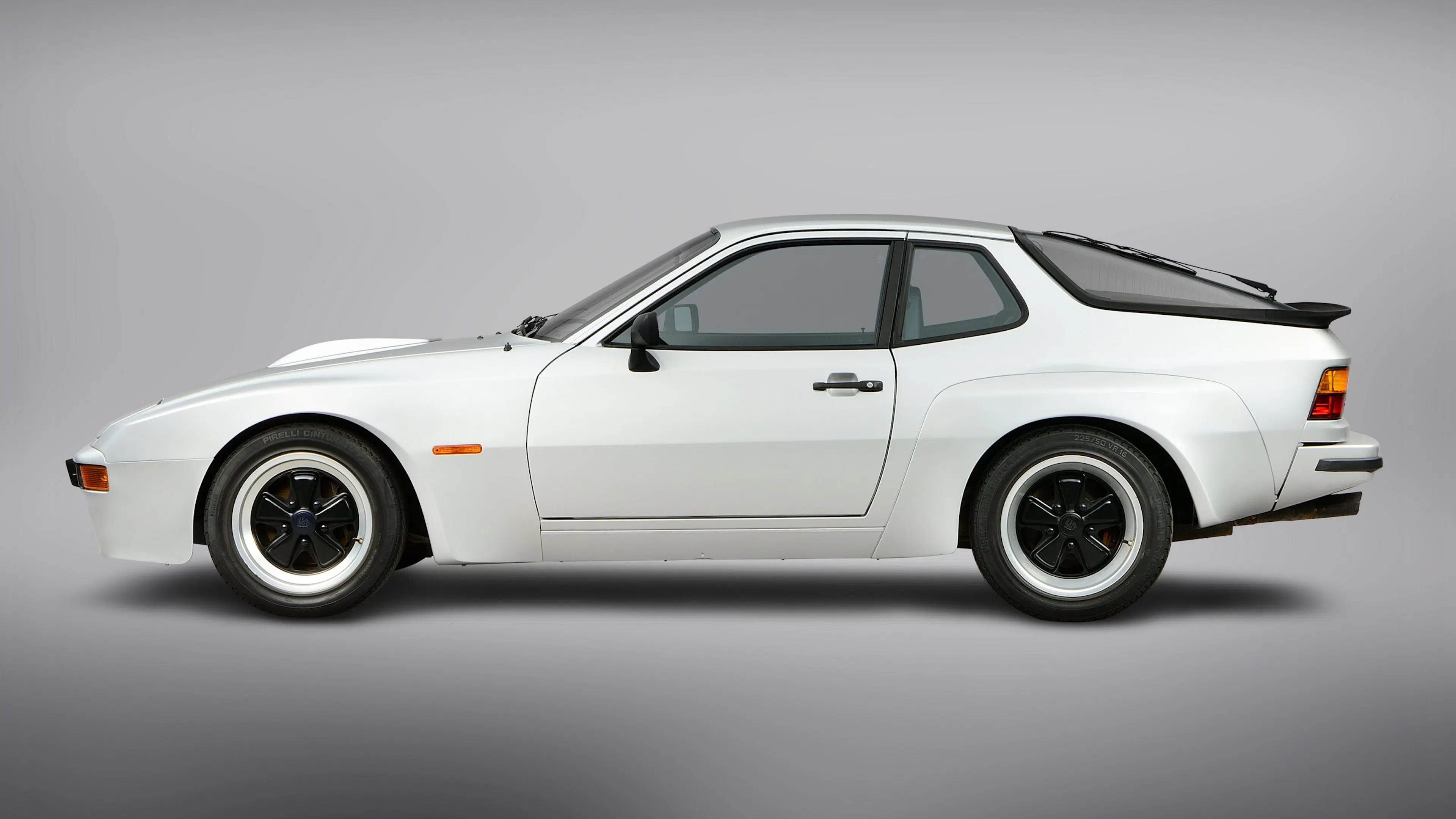 Weißer Porsche 924 steht seitlich vor neutralem, grauem Hintergrund, Klappscheinwerfer eingeklappt.