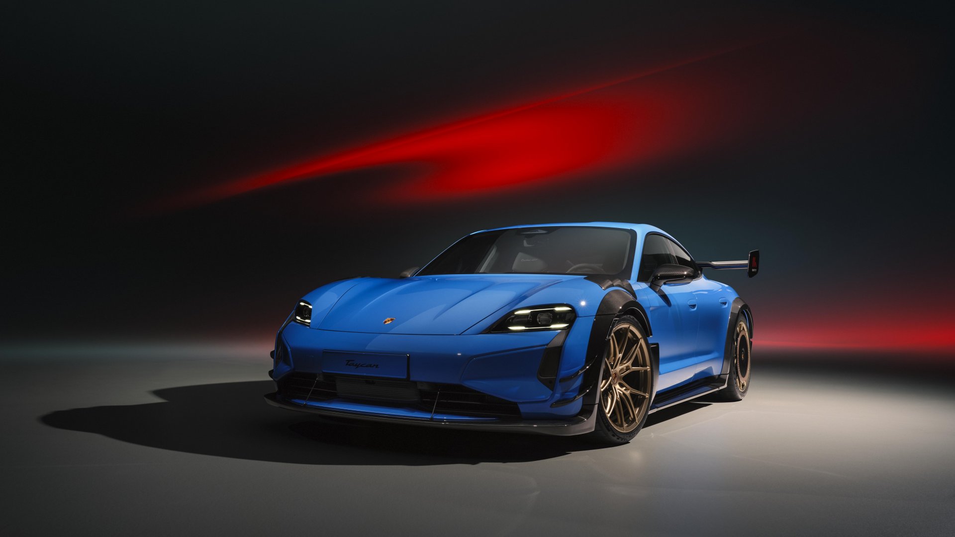 Porsche Taycan Turbo GT in Blau mit großem Heckflügel und bronzefarbenen Felgen, Studioaufnahme vor dunklem Hintergrund mit rotem Lichtstrahl.