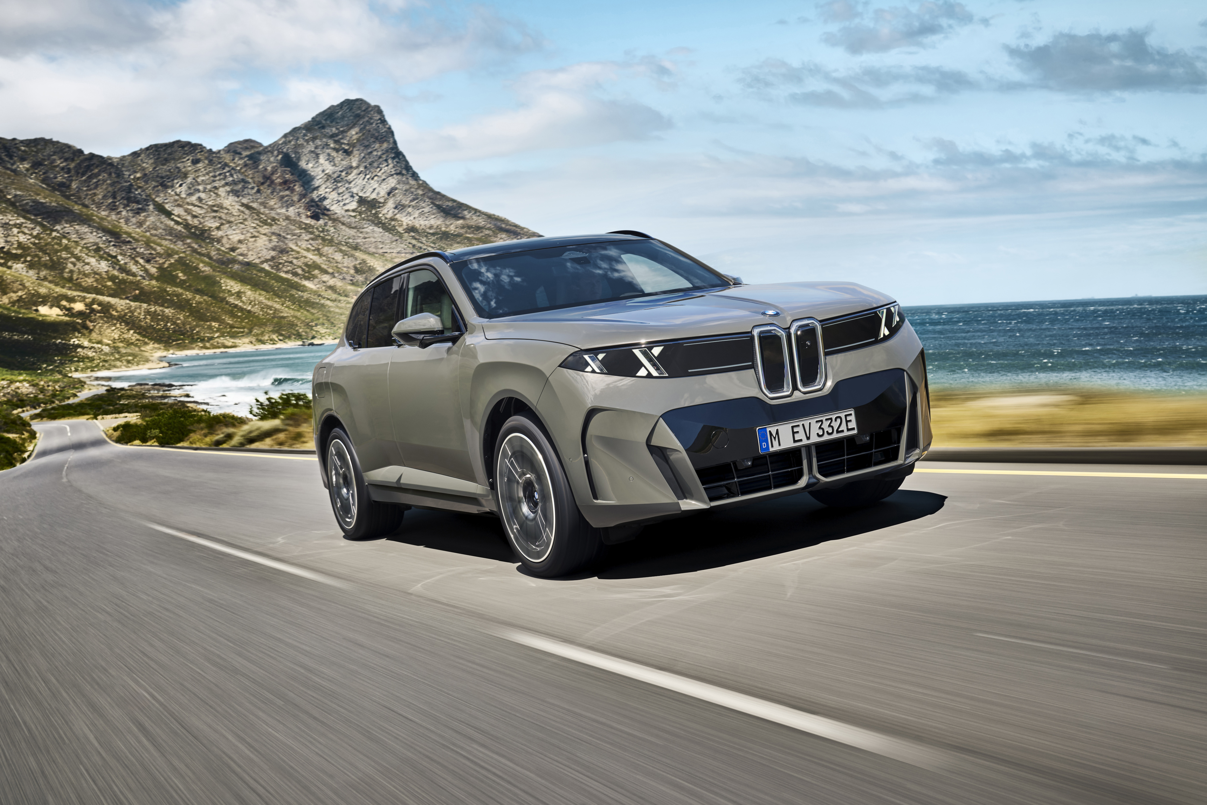 Graugrüner BMW iX5 fährt auf einer Küstenstraße, im Hintergrund Berge und Meer.