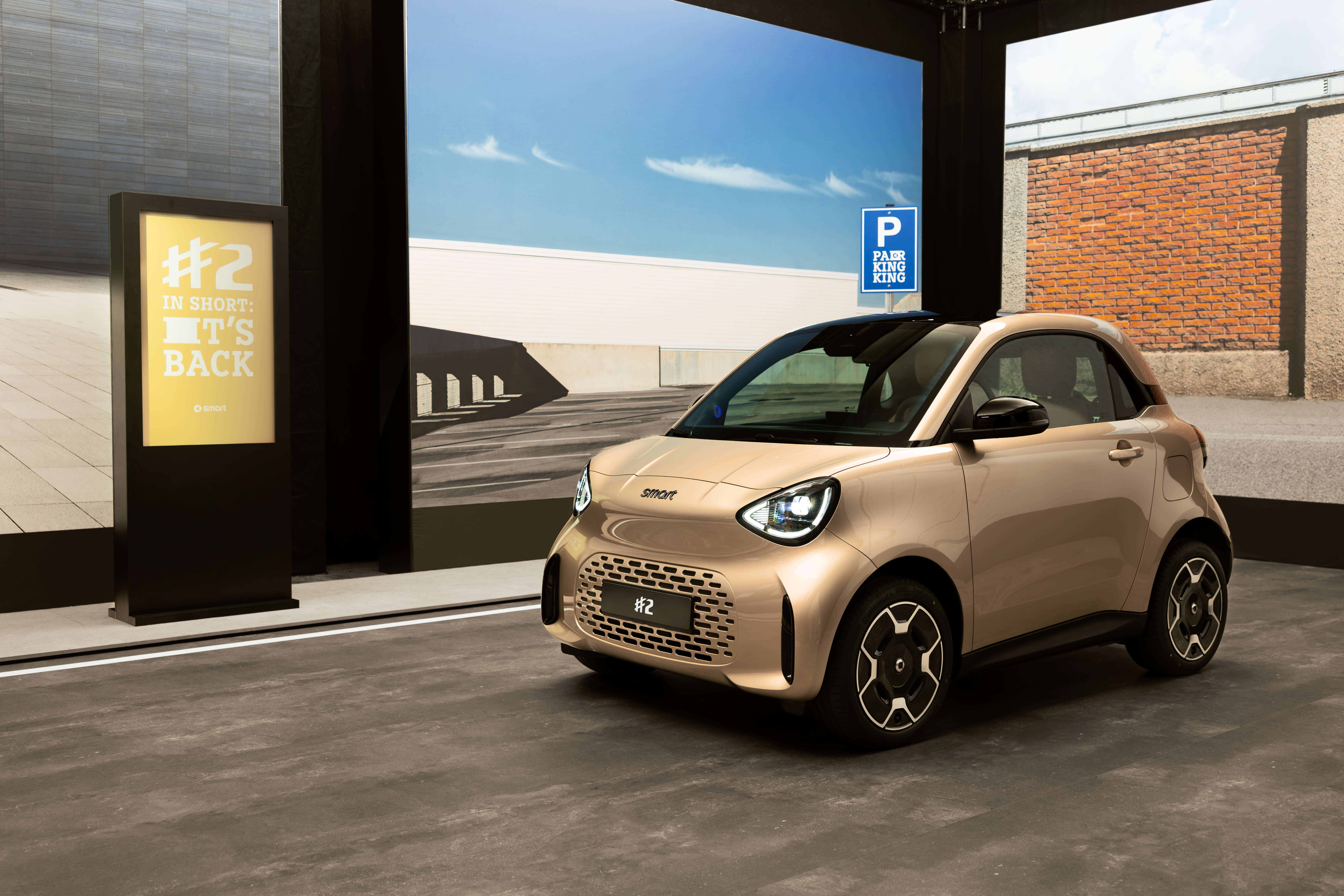 Champagnerfarbener smart #2 steht in einer Ausstellung neben einem Aufsteller mit der Aufschrift "It’s back" und einem Parkplatzschild.