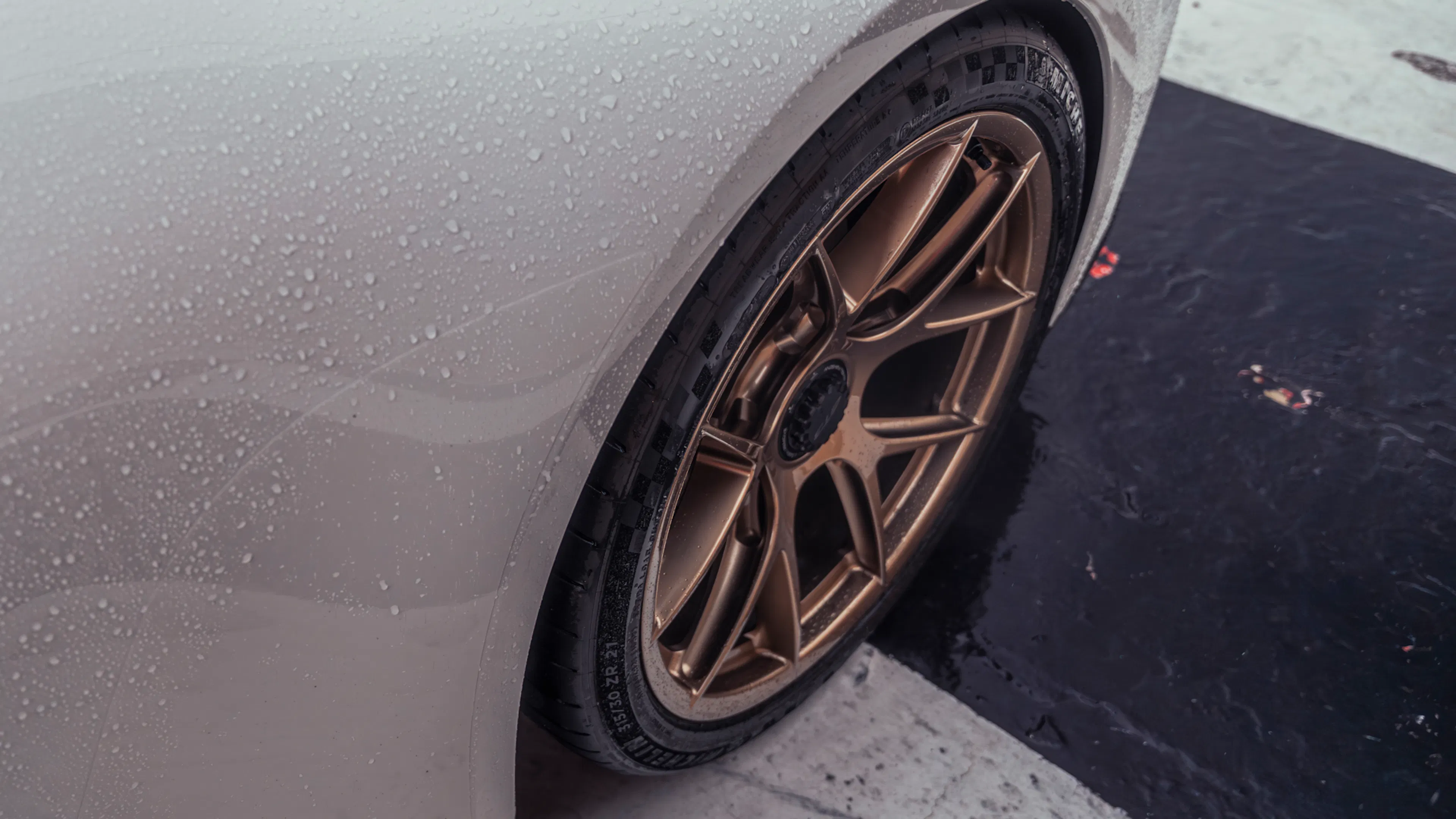 Nahaufnahme eines goldfarbenen Leichtmetallrads mit sportlichem Michelin-Reifen an einem weißen Porsche 911 GT3; Regentropfen bedecken den Kotflügel.