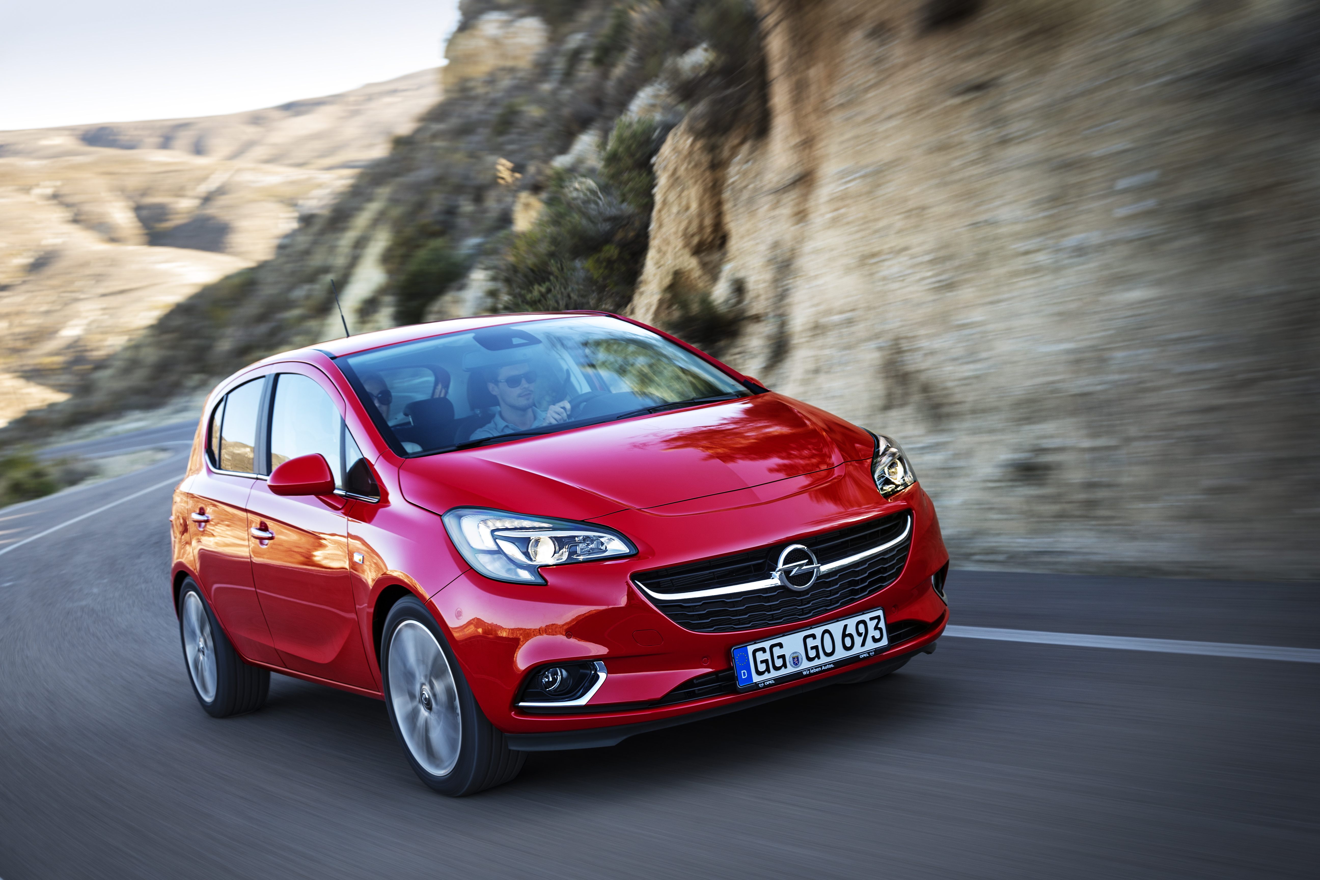 Roter Opel Corsa E fährt auf einer Landstraße an einer felsigen Böschung entlang. Zwei Personen sitzen im Wagen.