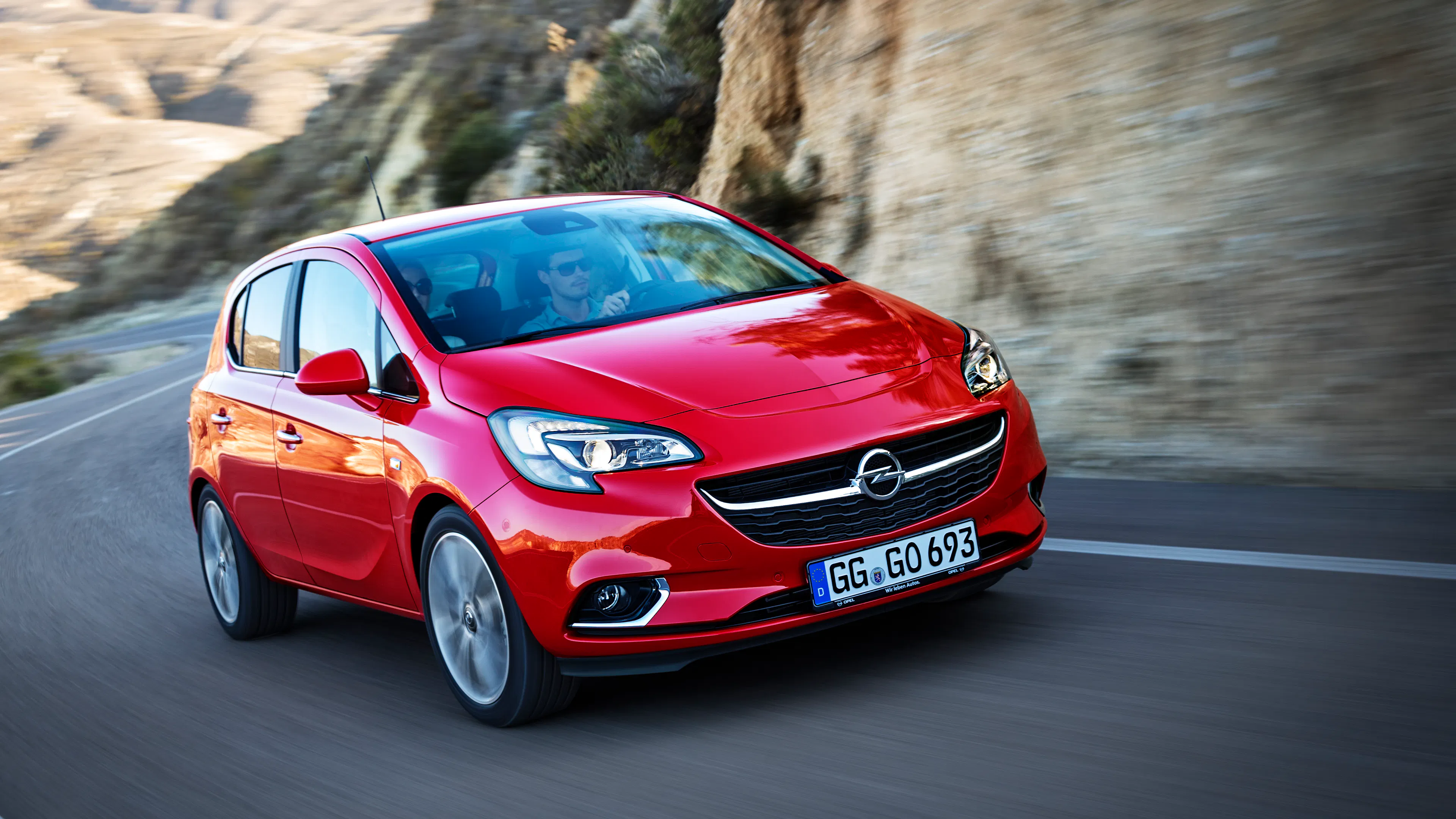 Roter Opel Corsa E fährt auf einer Landstraße an einer felsigen Böschung entlang. Zwei Personen sitzen im Wagen.