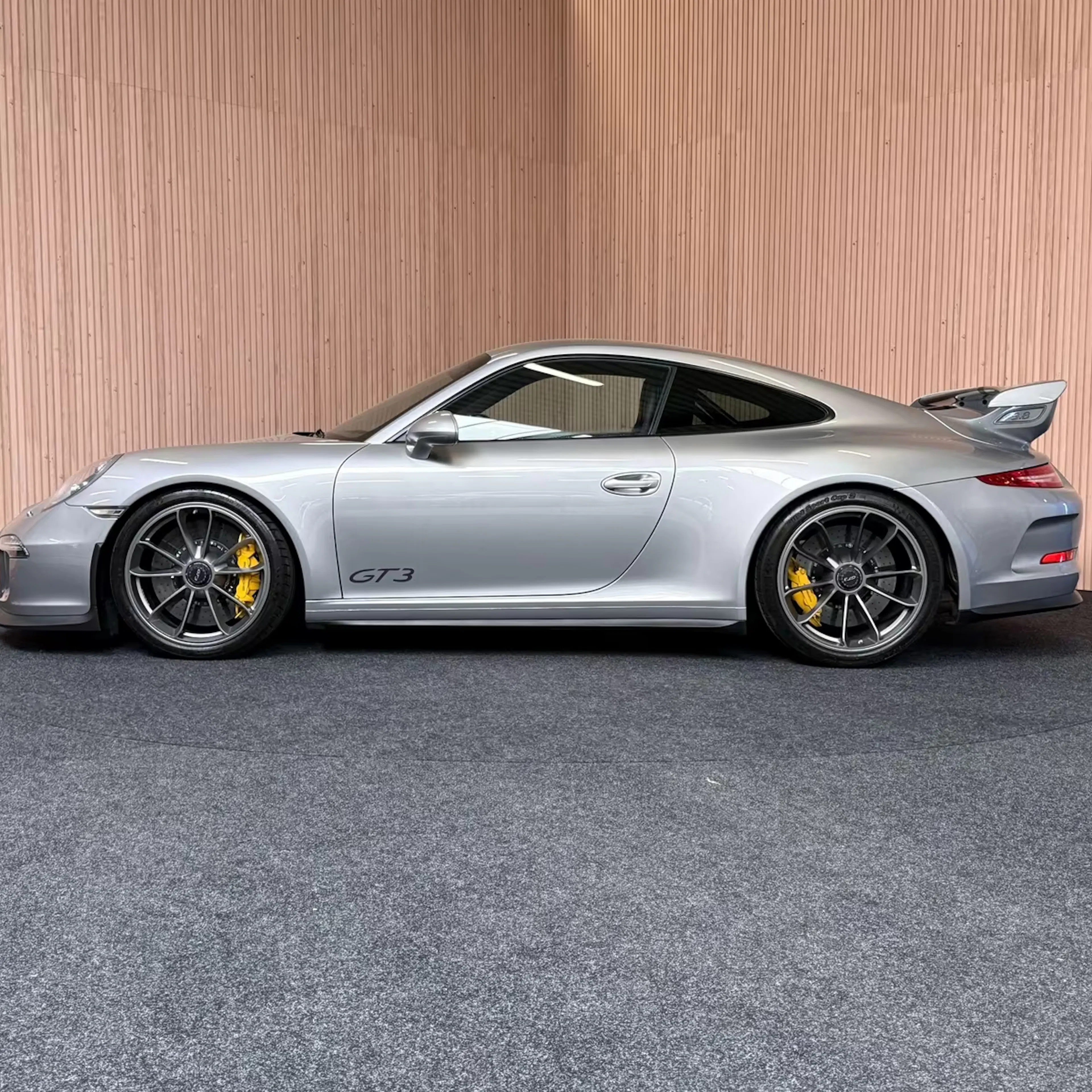 Silberner Porsche 911 GT3 steht vor Holzwand auf grauem Teppich