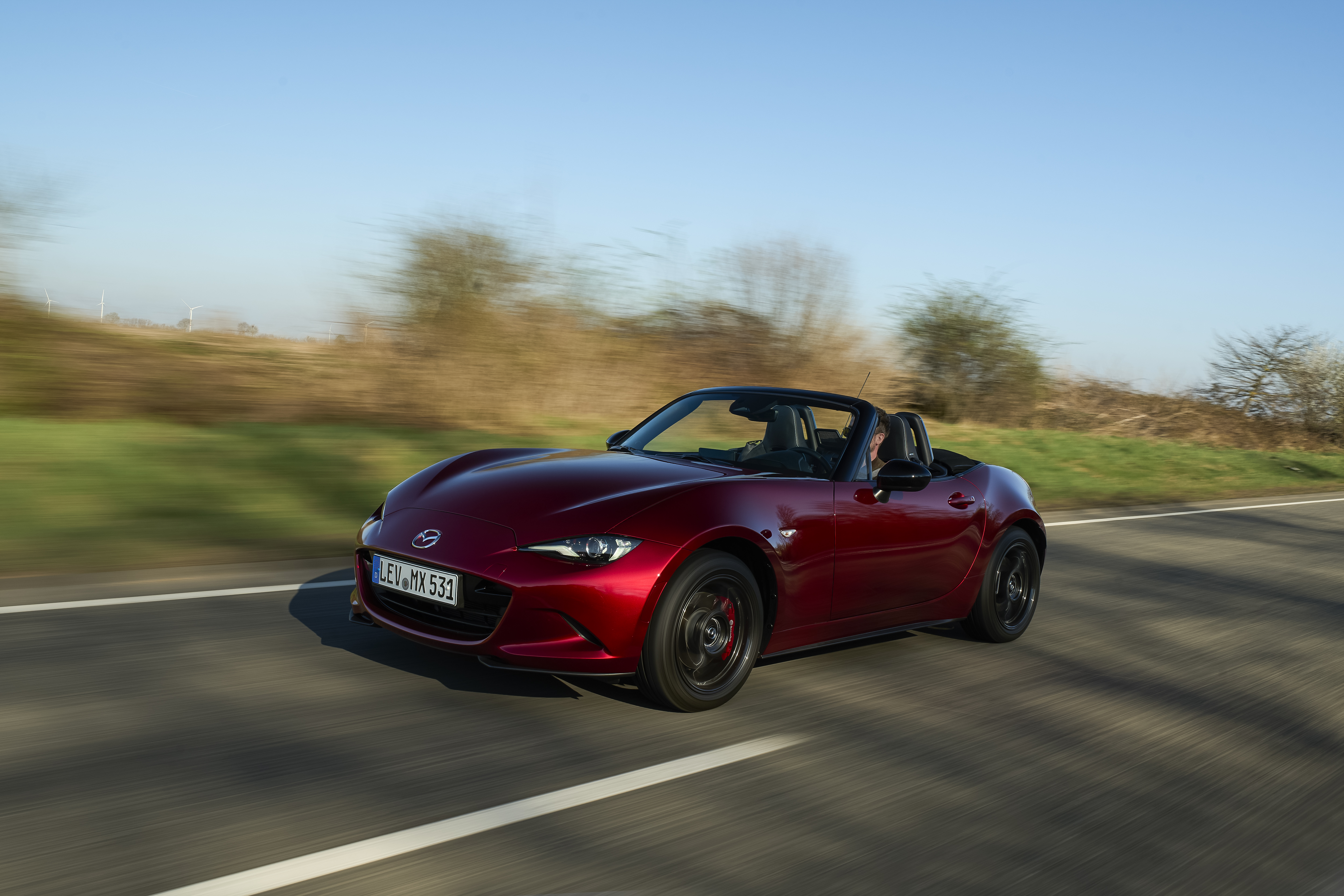 Mazda MX-5 2027 fährt dynamisch auf einer Landstraße, Seitenansicht von vorne, Verdeck geöffnet