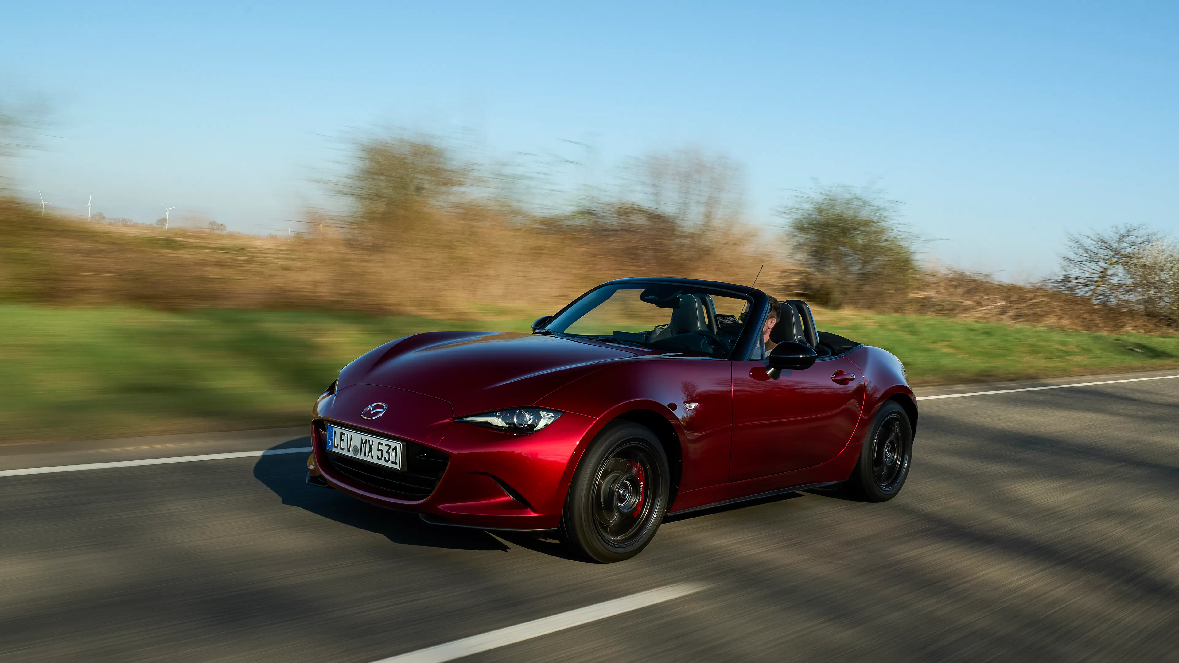 Mazda MX-5 2027 fährt dynamisch auf einer Landstraße, Seitenansicht von vorne, Verdeck geöffnet