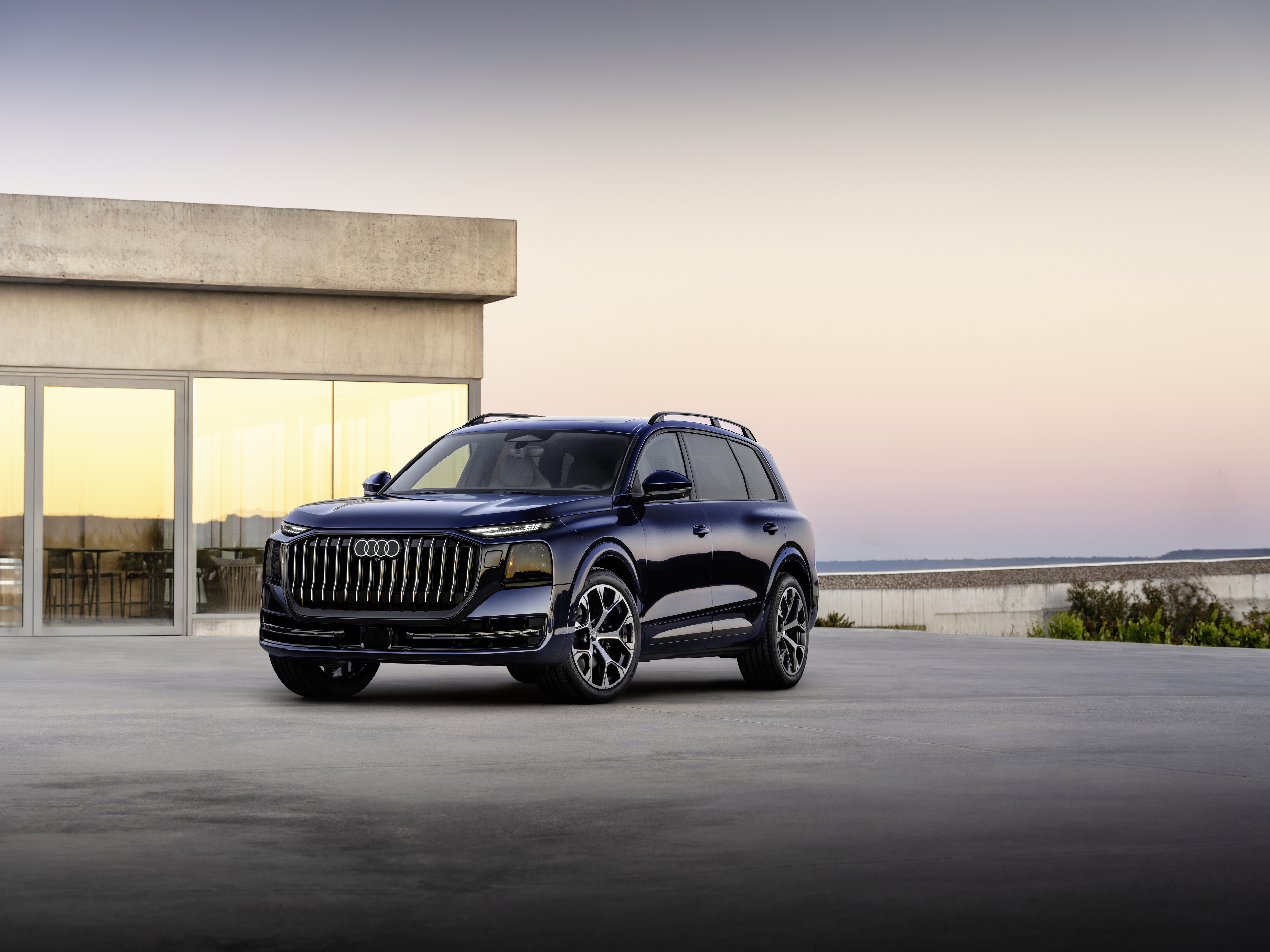 Audi Q9 in Dunkelblau seitlich vor einem modernen Betongebäude bei Dämmerung.