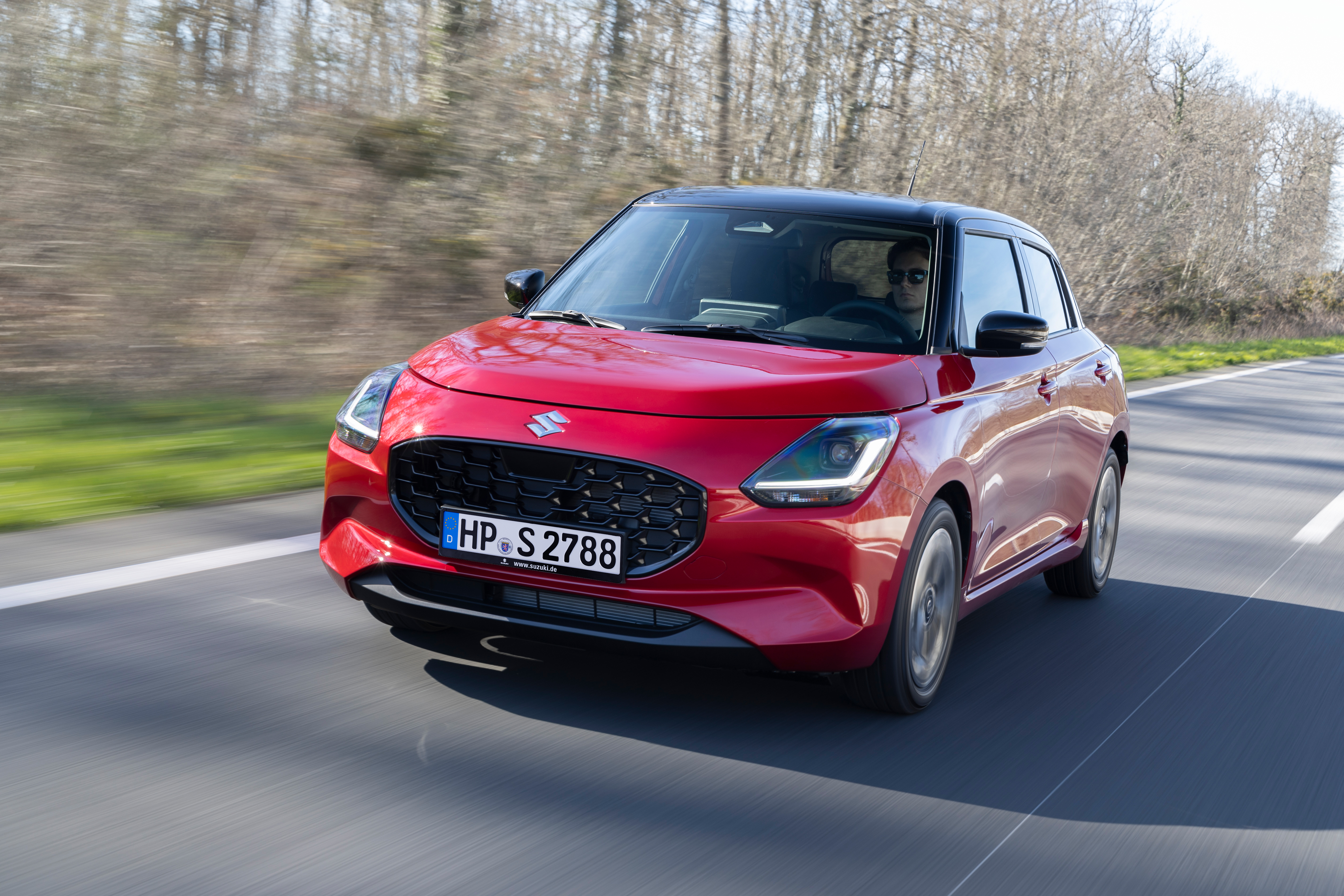 Roter Suzuki Swift mit schwarzem Dach fährt auf einer Landstraße, im Hintergrund kahle Bäume.