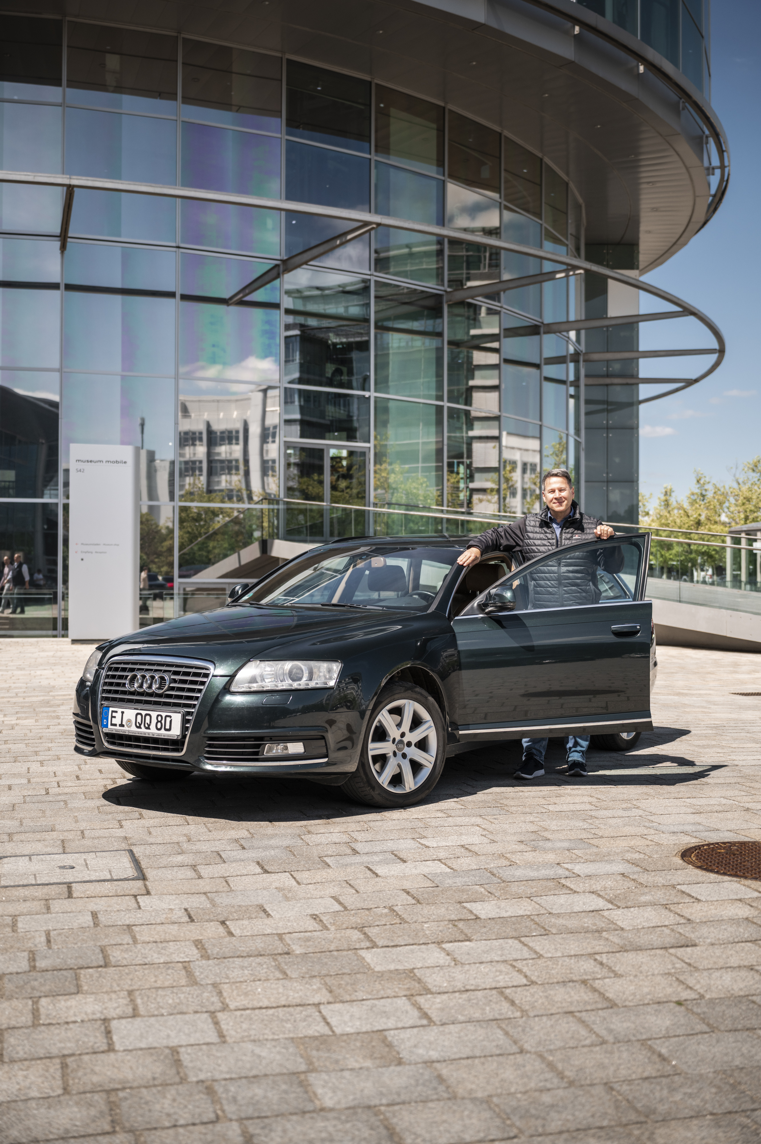 Dunkelgrüner Audi A6 C6 mit geöffneter Fahrertür vor dem Audi museum mobile, Besitzer steht daneben.