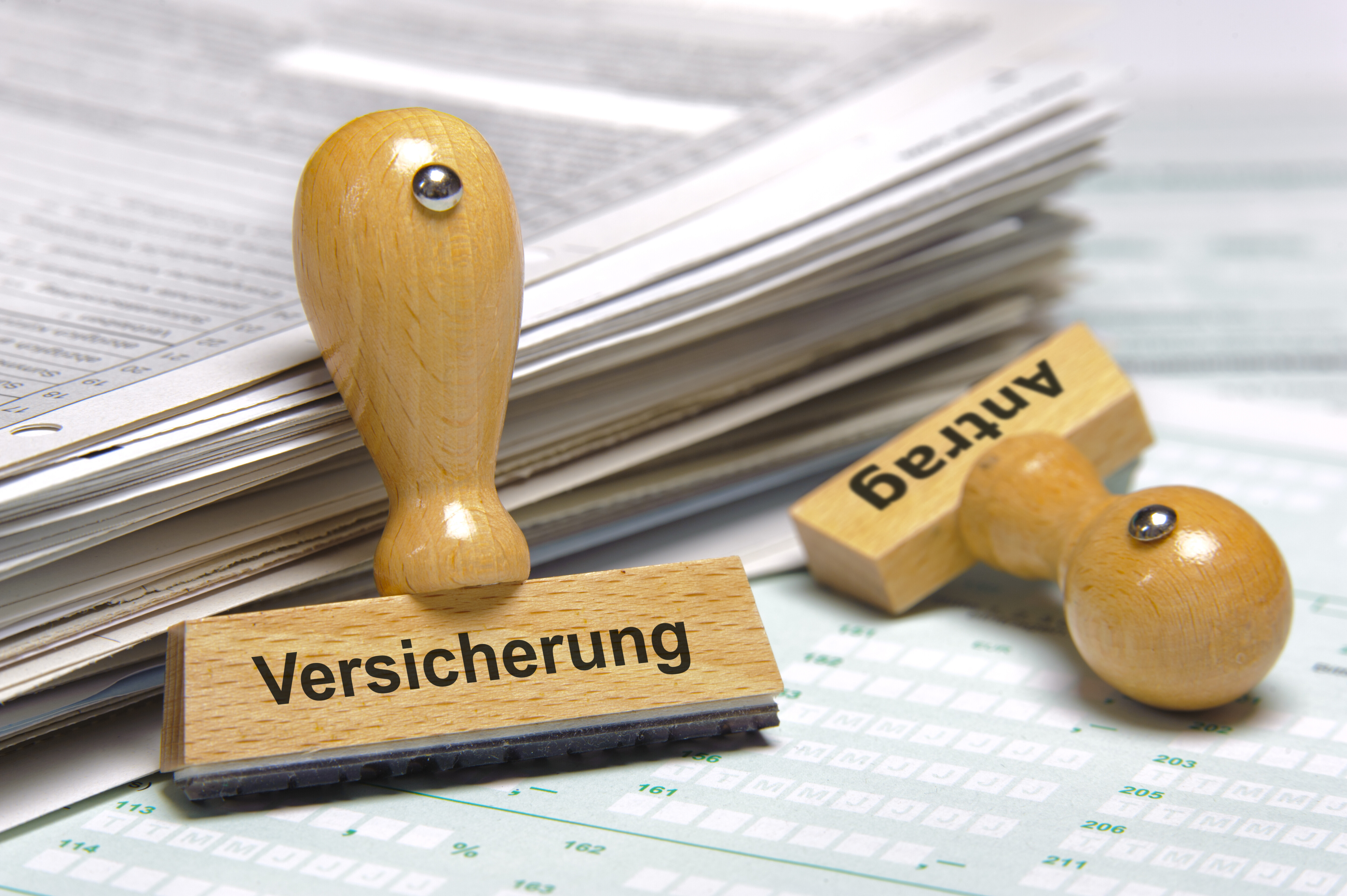 Zwei Stempel liegen neben einem Papierstapel. Auf einem steht "Versicherung", auf dem anderen "Antrag".