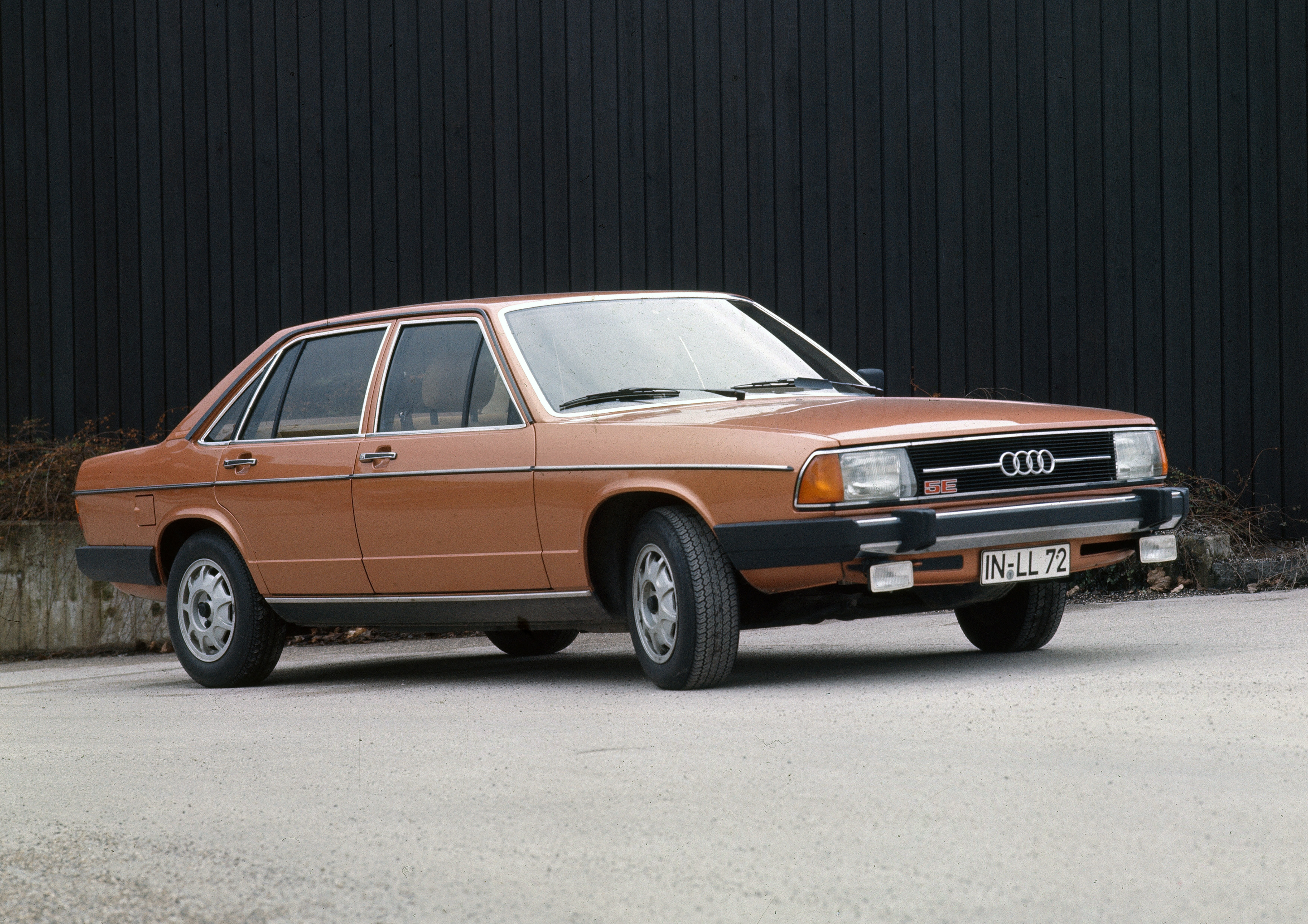 Kupferfarbener Audi 100 C2 der zweiten Generation, seitlich fotografiert vor einer dunklen Holzwand.