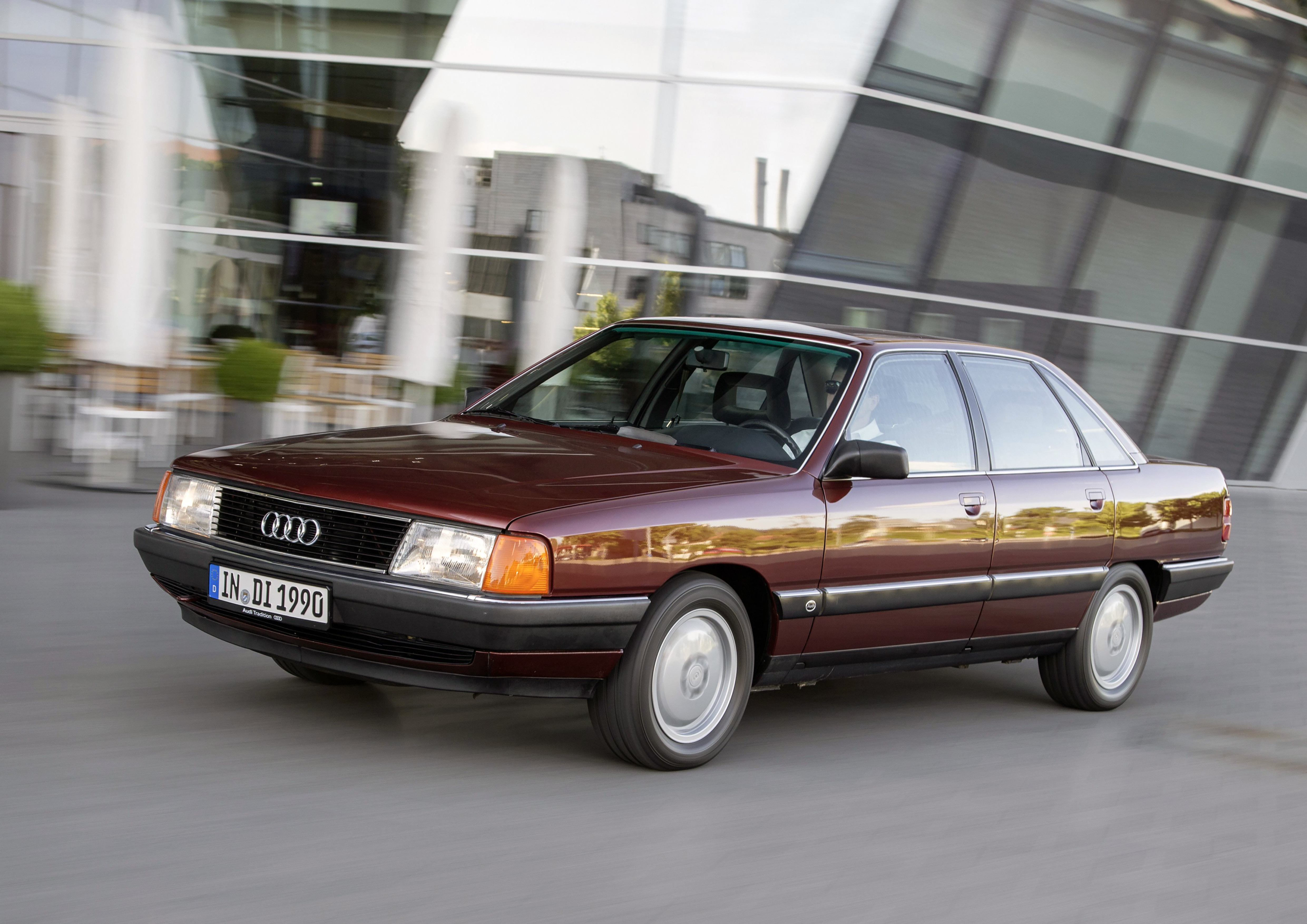 Dunkelroter Audi 100 C3 in Fahrt vor einer modernen Glasfassade, aufgenommen von der Seite schräg vorn.