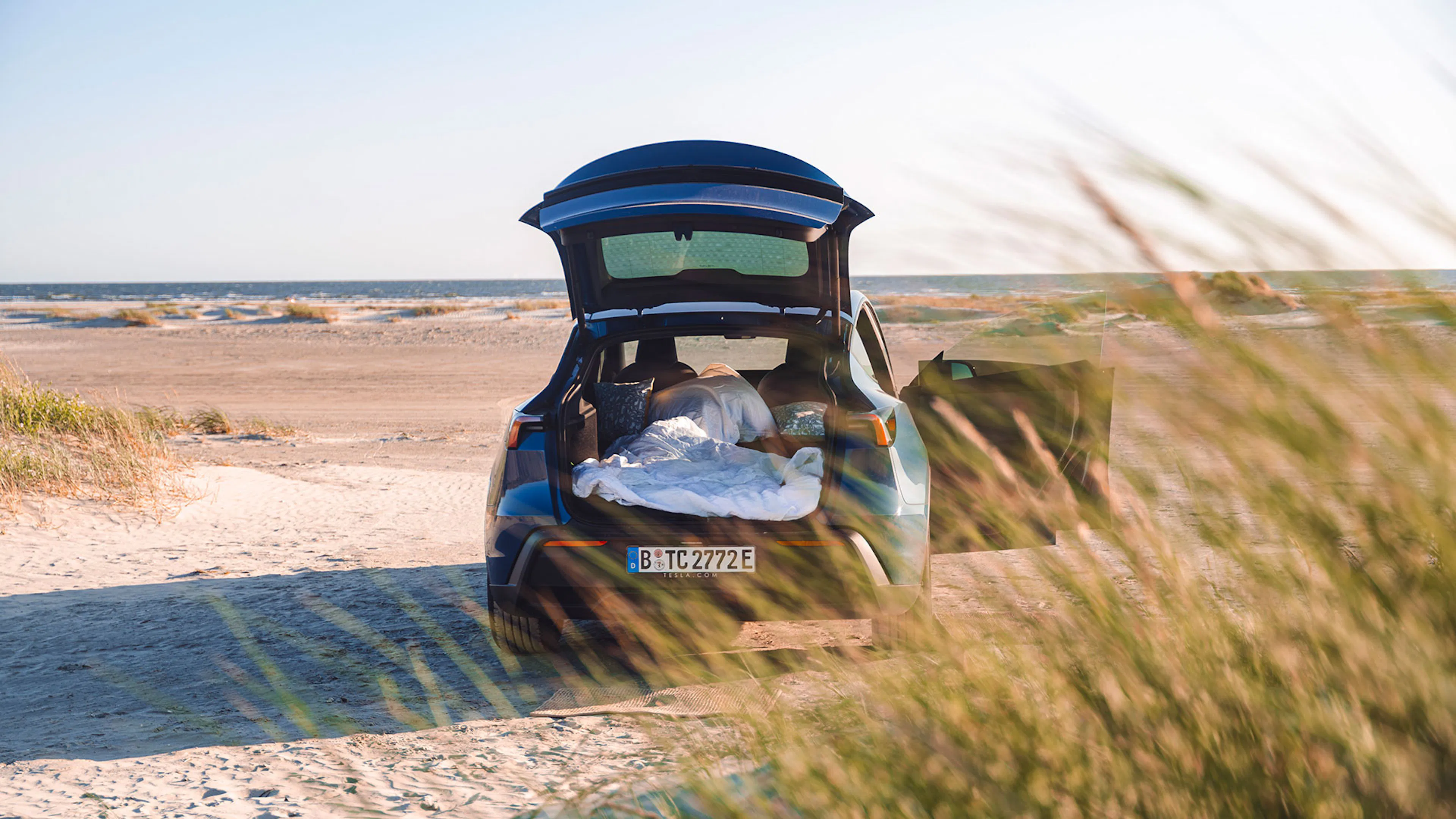 Ein blaues Tesla Model Y steht mit geöffnetem Kofferraum und Beifahrertür am Strand, die Luftmatratze und Decken fürs Campen vorbereitet.