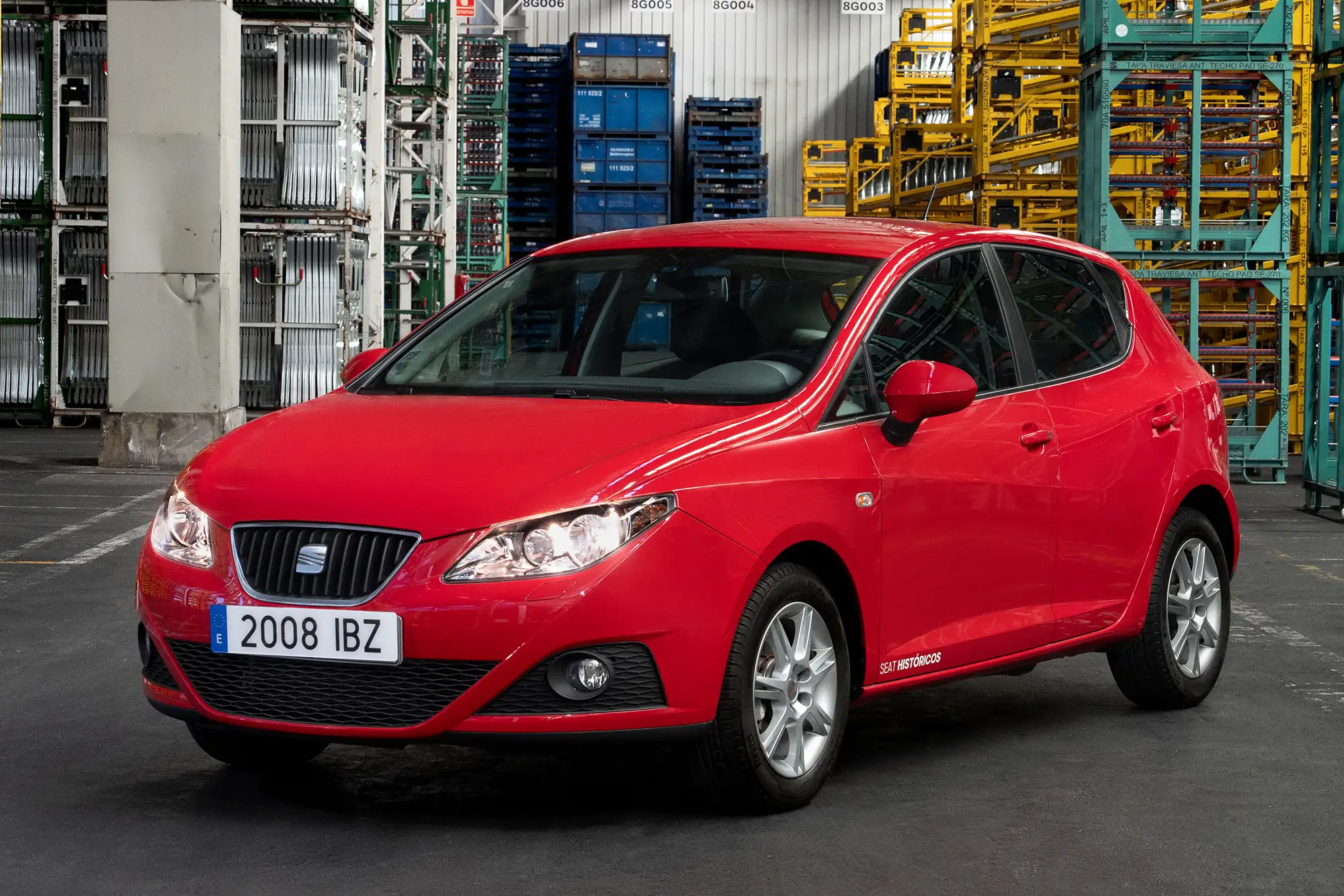 Roter Seat Ibiza mit eingeschalteten Scheinwerfern steht vor Gitterboxen und Regalen in einer Werkshalle.