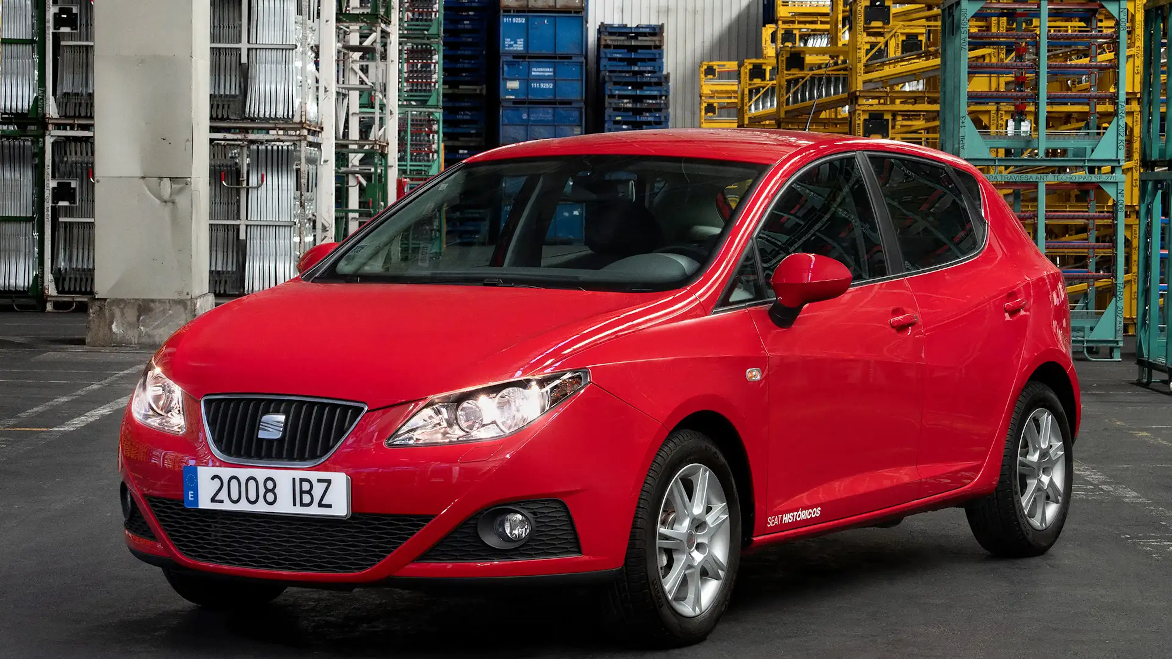 Roter Seat Ibiza mit eingeschalteten Scheinwerfern steht vor Gitterboxen und Regalen in einer Werkshalle.