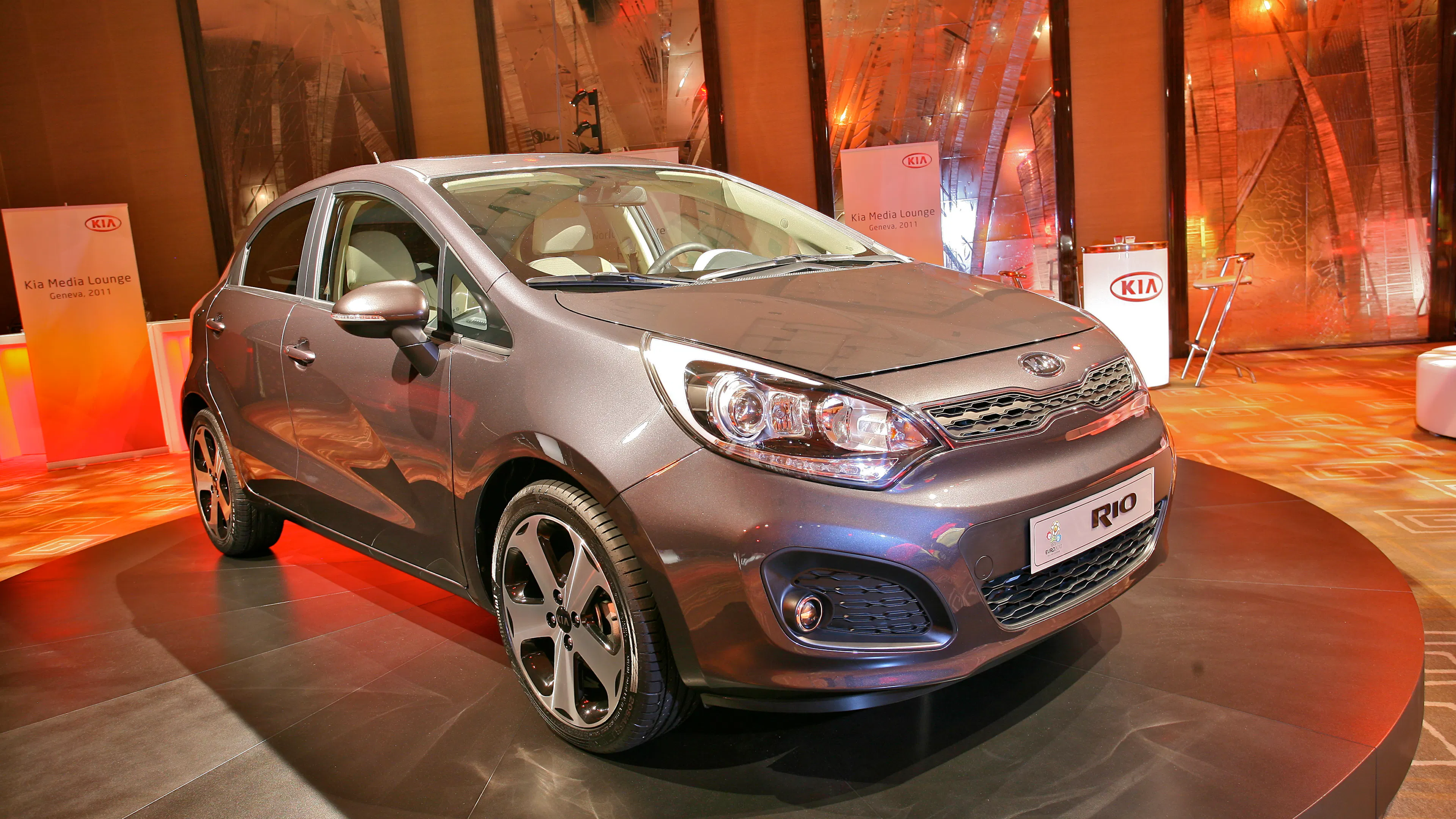 Graubrauner Kia Rio III steht auf einem runden, schwarzen Podest in einer orange beleuchteten Messelounge.