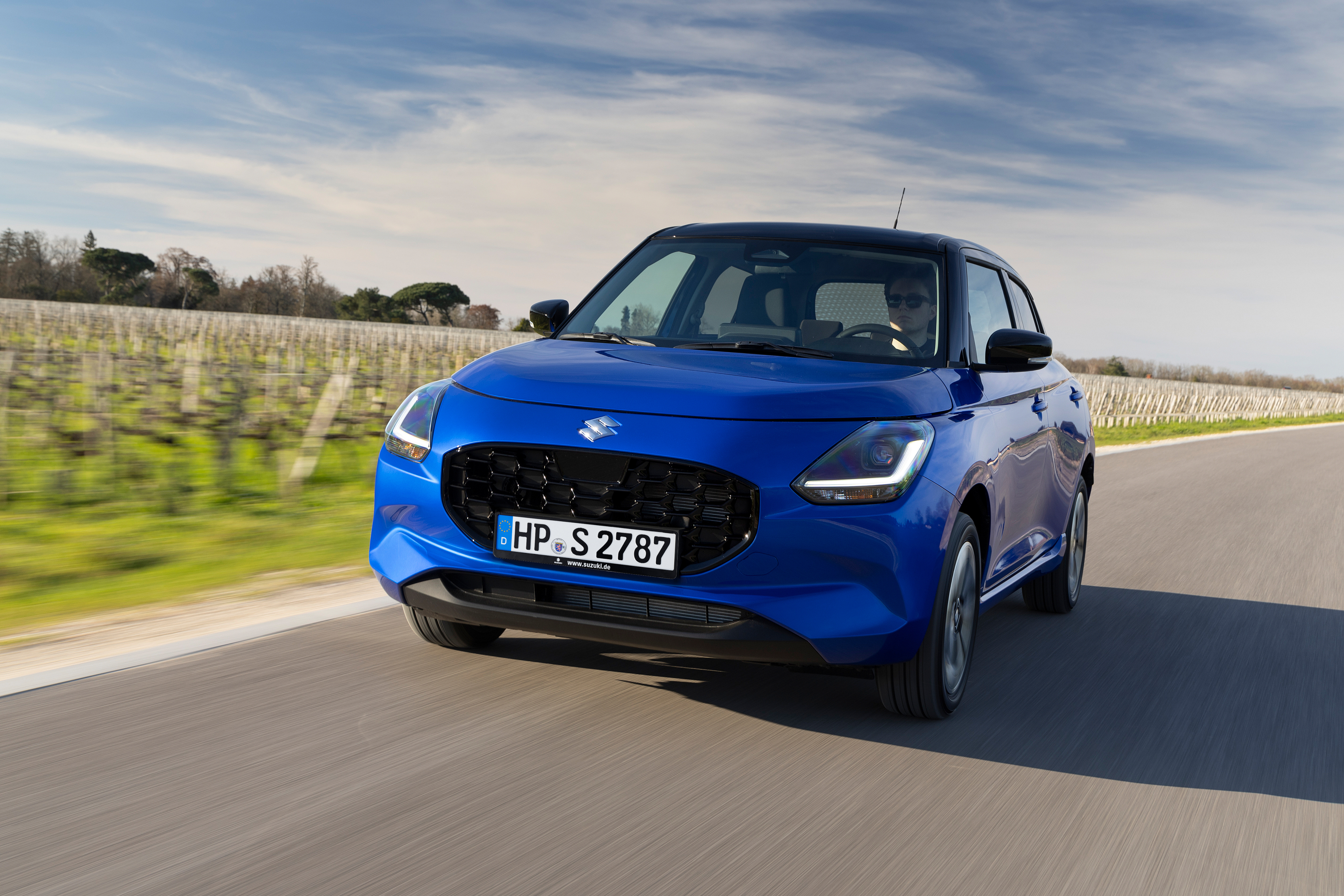 Blauer Suzuki Swift mit schwarzem Dach fährt frontal auf einer Landstraße neben einem Weinberg.