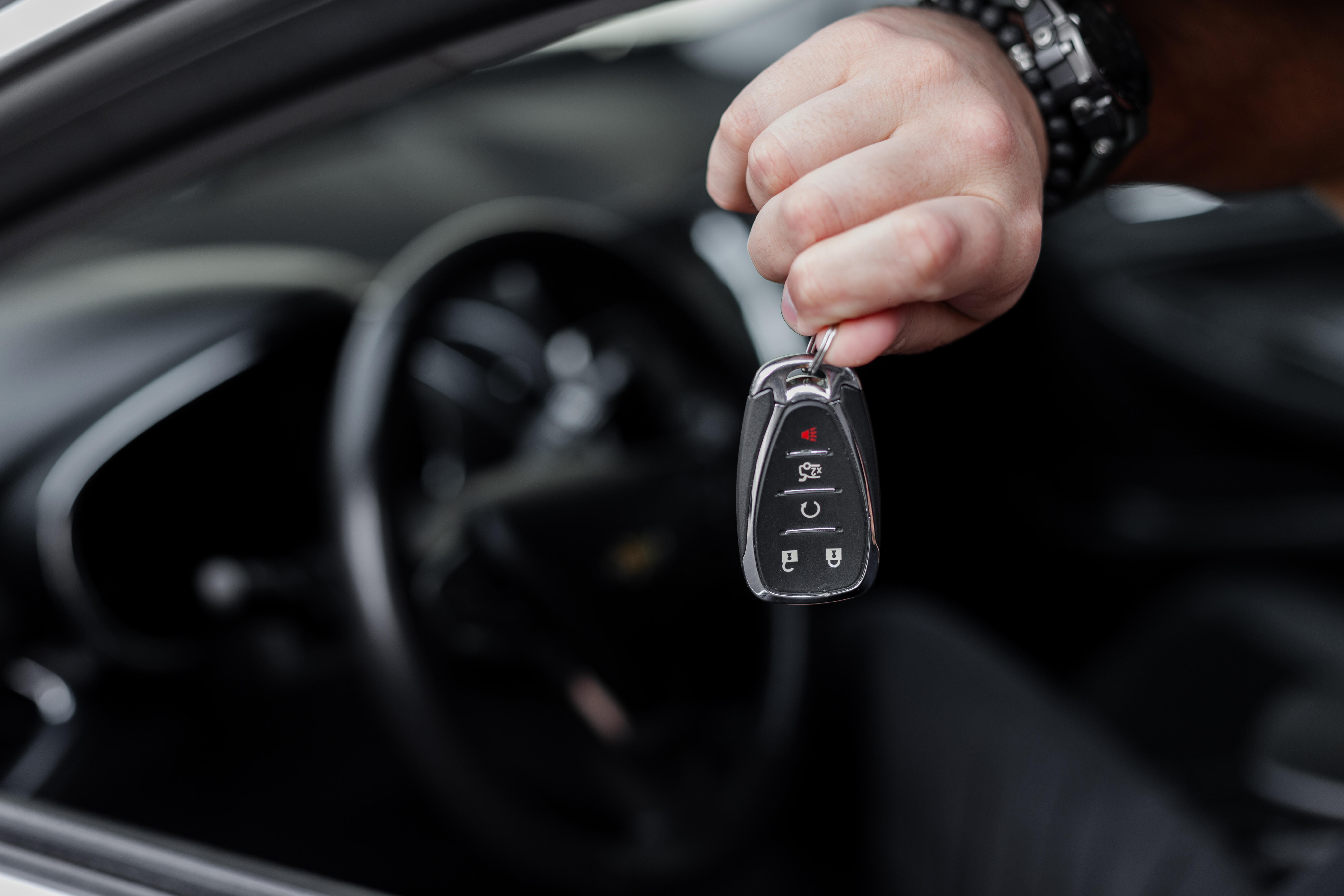 Hand hält einen Funkschlüssel mit mehreren Tasten vor dem Lenkrad eines Autos, der Innenraum ist unscharf zu sehen.