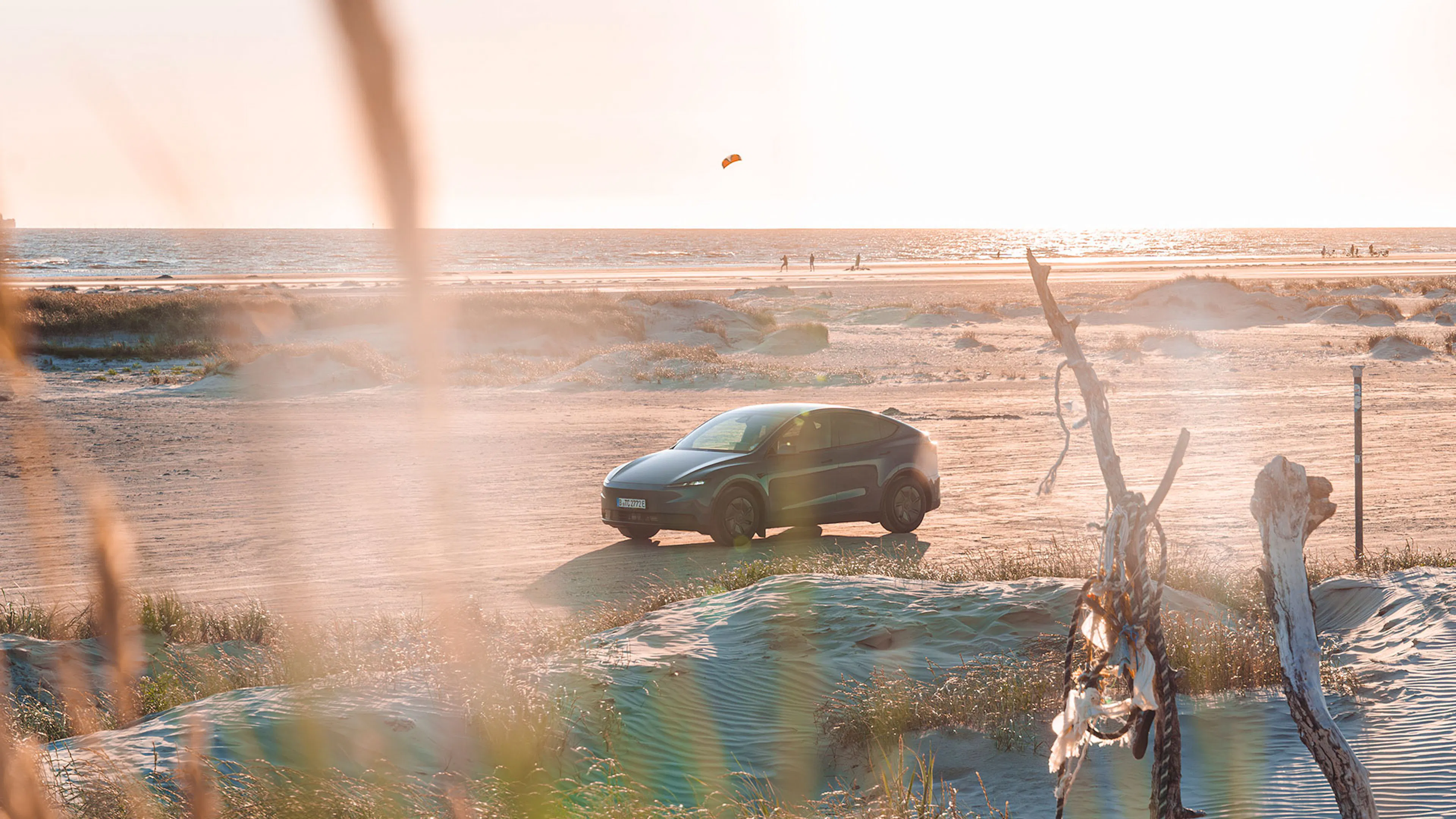 Ein hellblaues Tesla Model Y steht an einem Strand, die Sonne geht unter, im Hintergrund sind das Meer und ein Windsurfer.