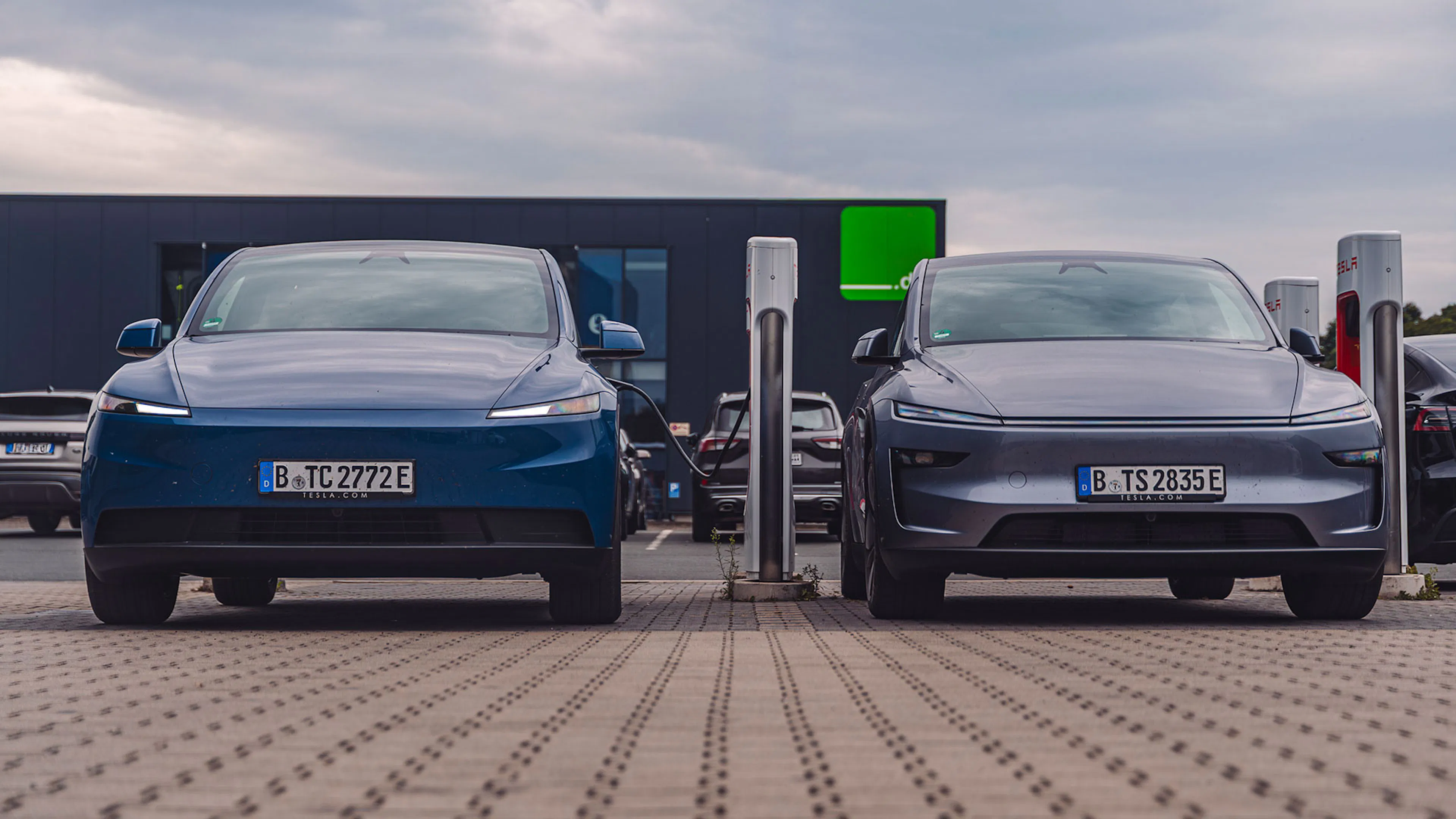 Ein blaues und ein graues Tesla Model Y stehen an einer Ladestation nebeneinander, Frontansicht.