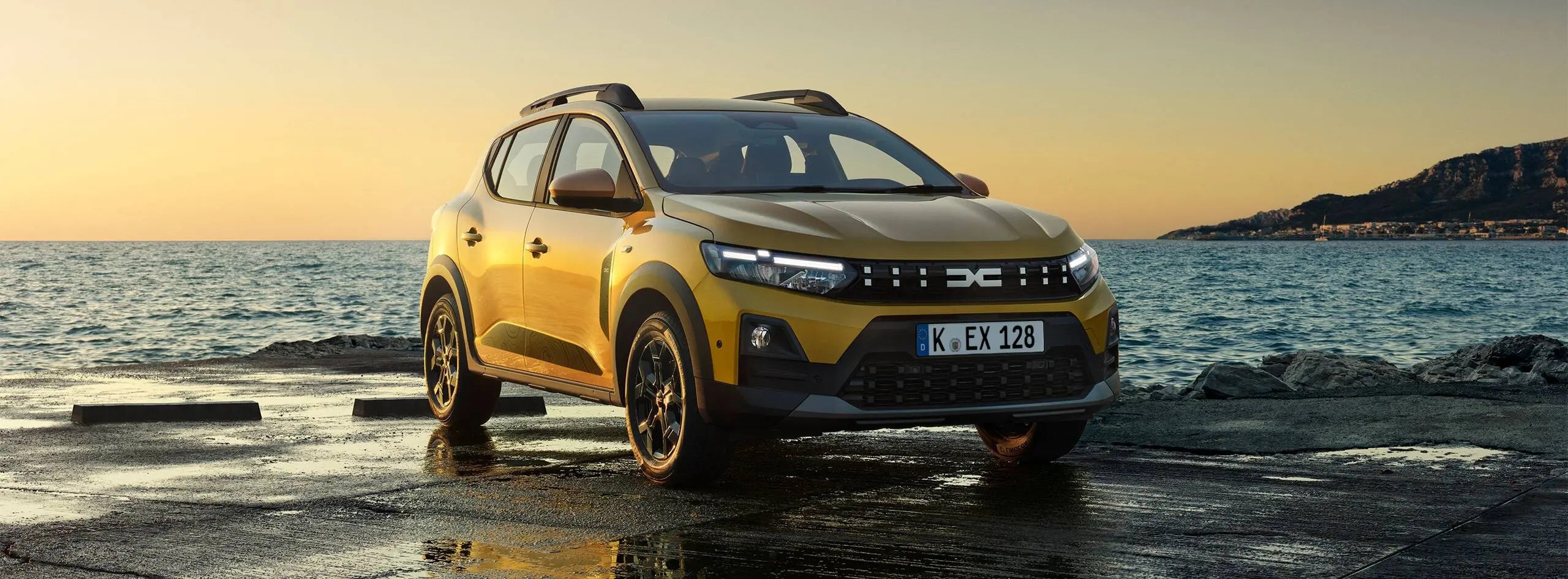 Goldgelber Dacia Sandero Stepway Hybrid 155 mit Dachreling auf einer nassen Kaimauer am Meer bei Sonnenuntergang.