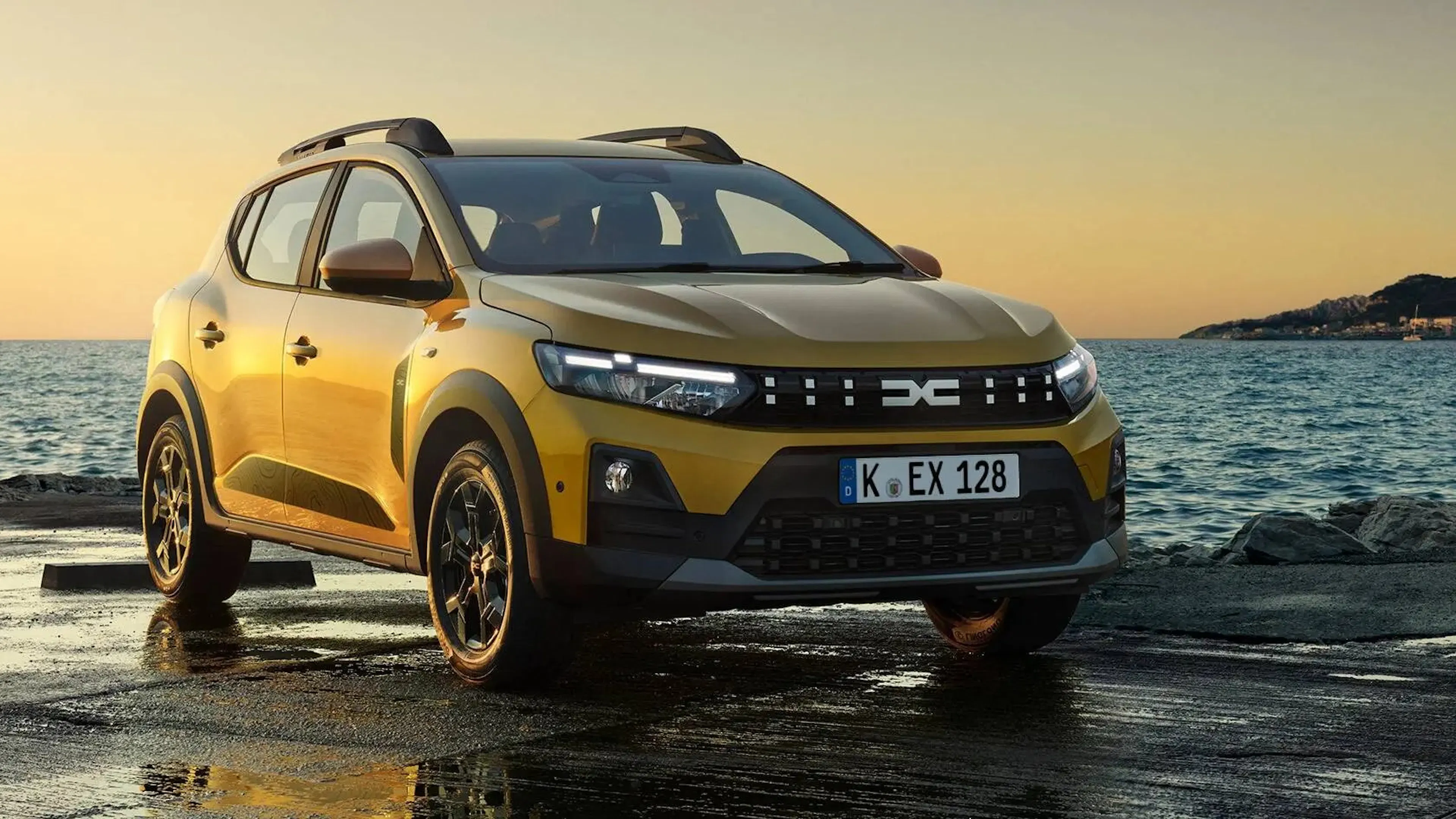 Goldgelber Dacia Sandero Stepway Hybrid 155 mit Dachreling auf einer nassen Kaimauer am Meer bei Sonnenuntergang.