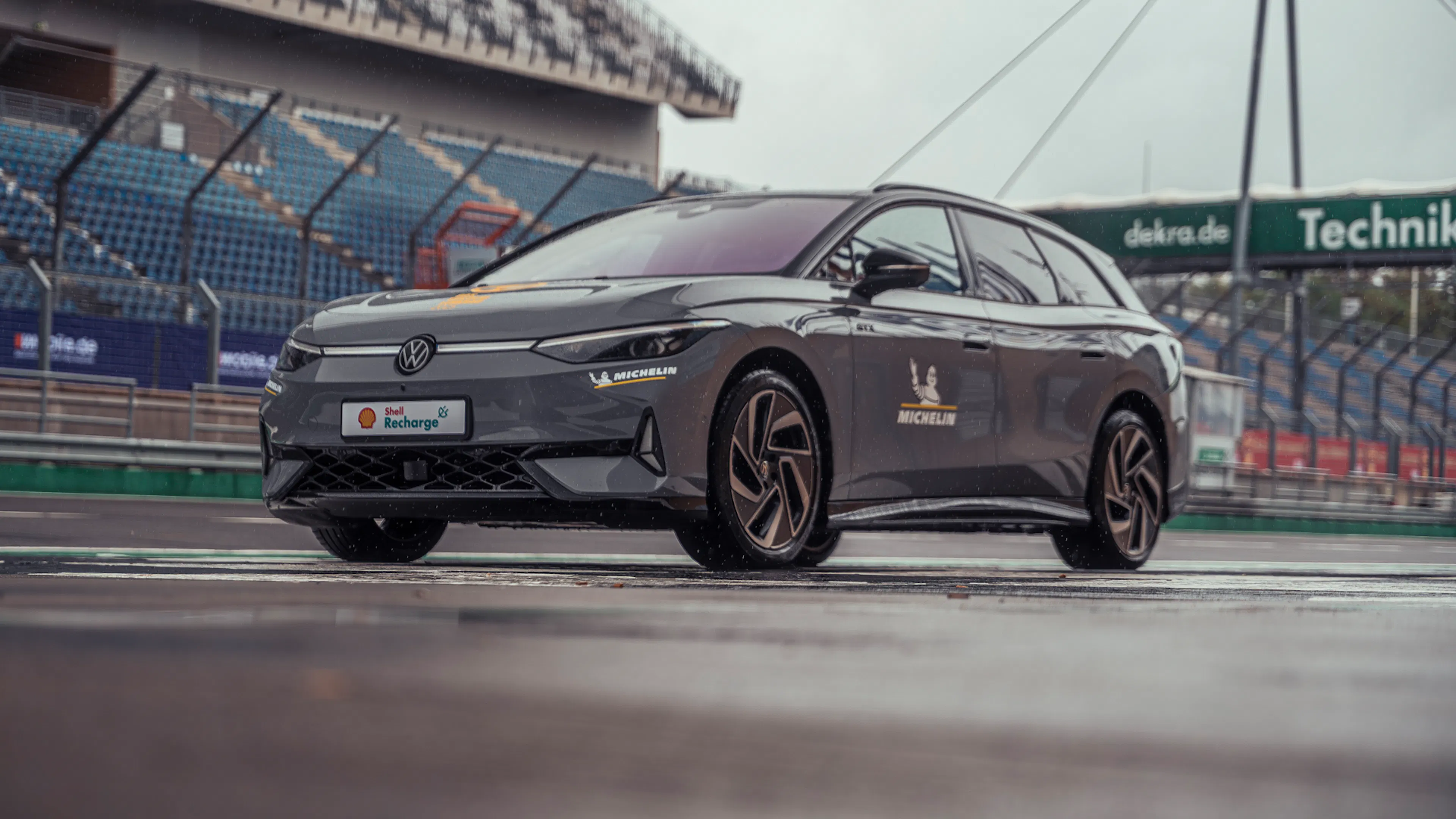 Grauer VW ID.7 Tourer GTX auf nasser Rennstrecke vor Tribünen und Boxengebäuden.